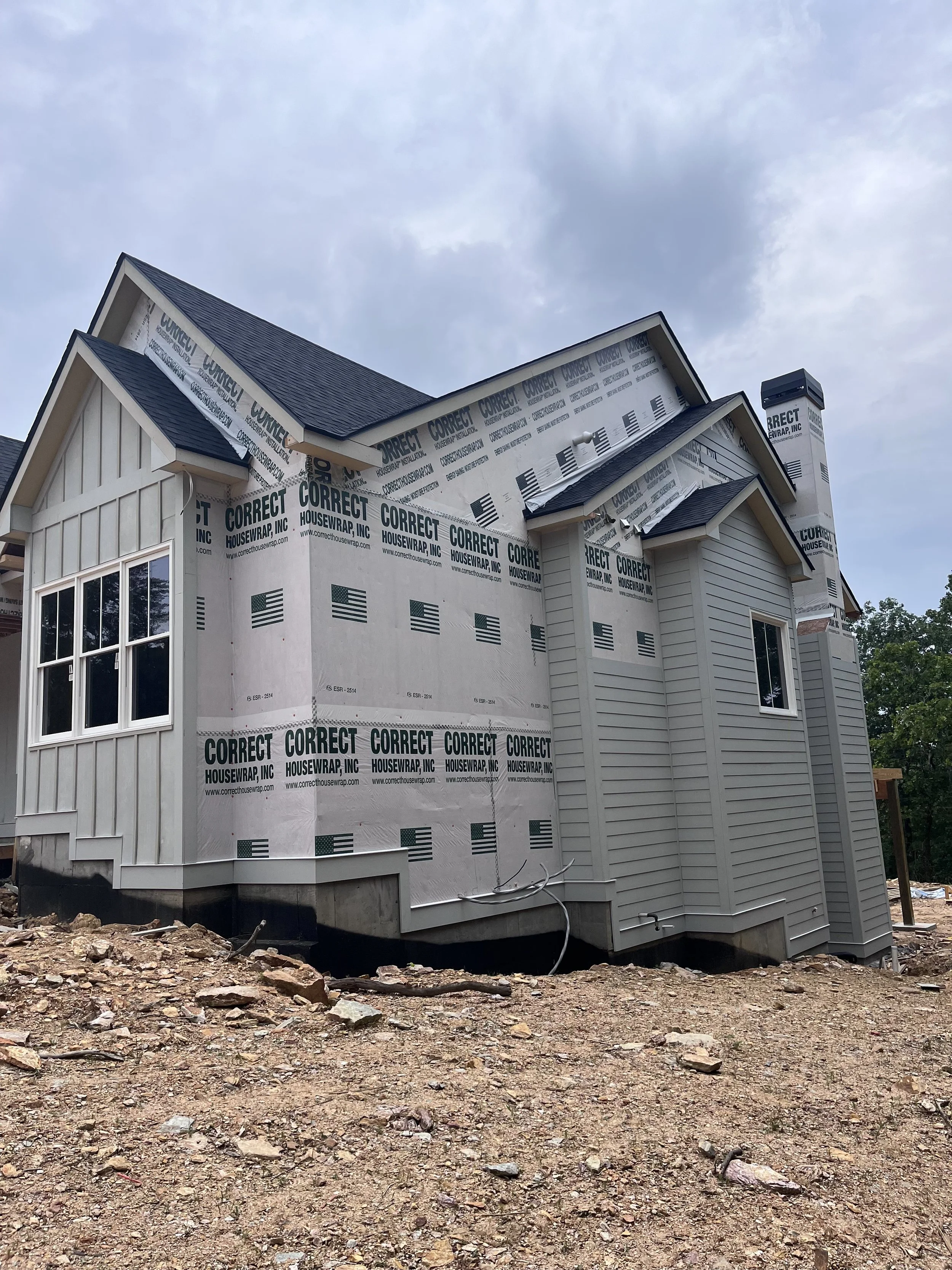 A house under construction with completed siding on one side and house wrap on the other, featuring a steep gable roof, a chimney, and a window, on a dirt lot with scattered rocks and debris.