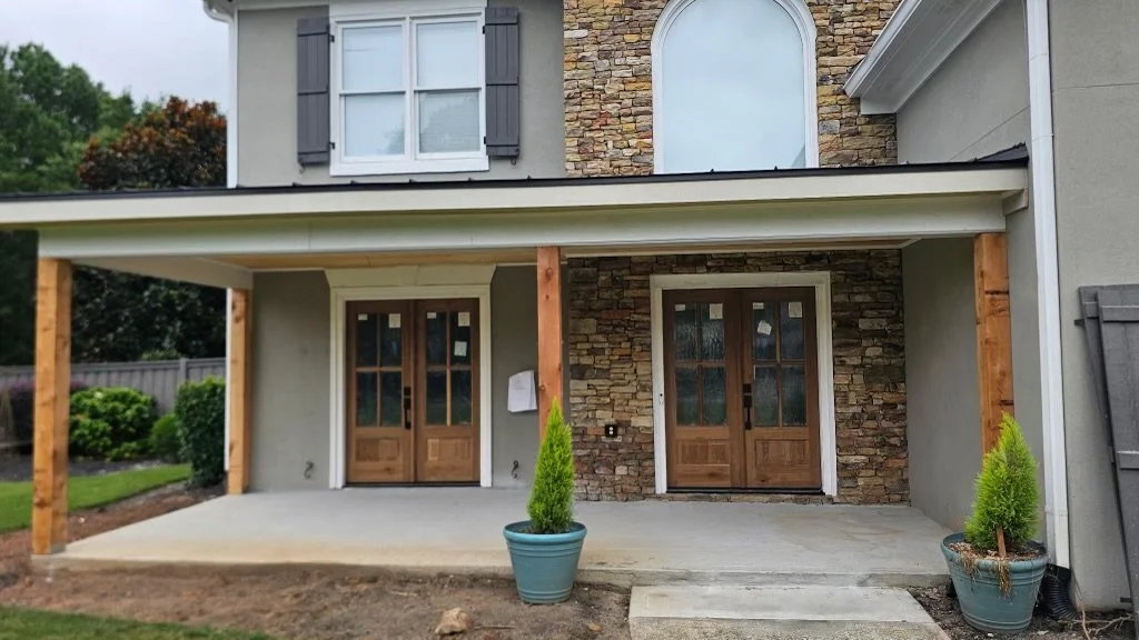 A house with a stone wall facade and wooden double doors, with a patio area in front, two potted plants, and a covered overhang supported by wooden posts.