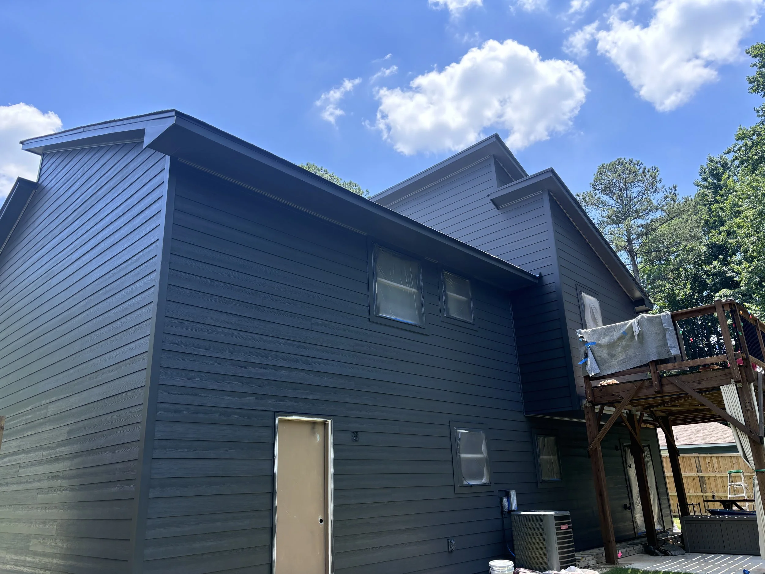 A two-story house with blue siding, rectangular windows, and an outdoor laundry area with a wooden balcony, ladder, and some cleaning supplies underneath.