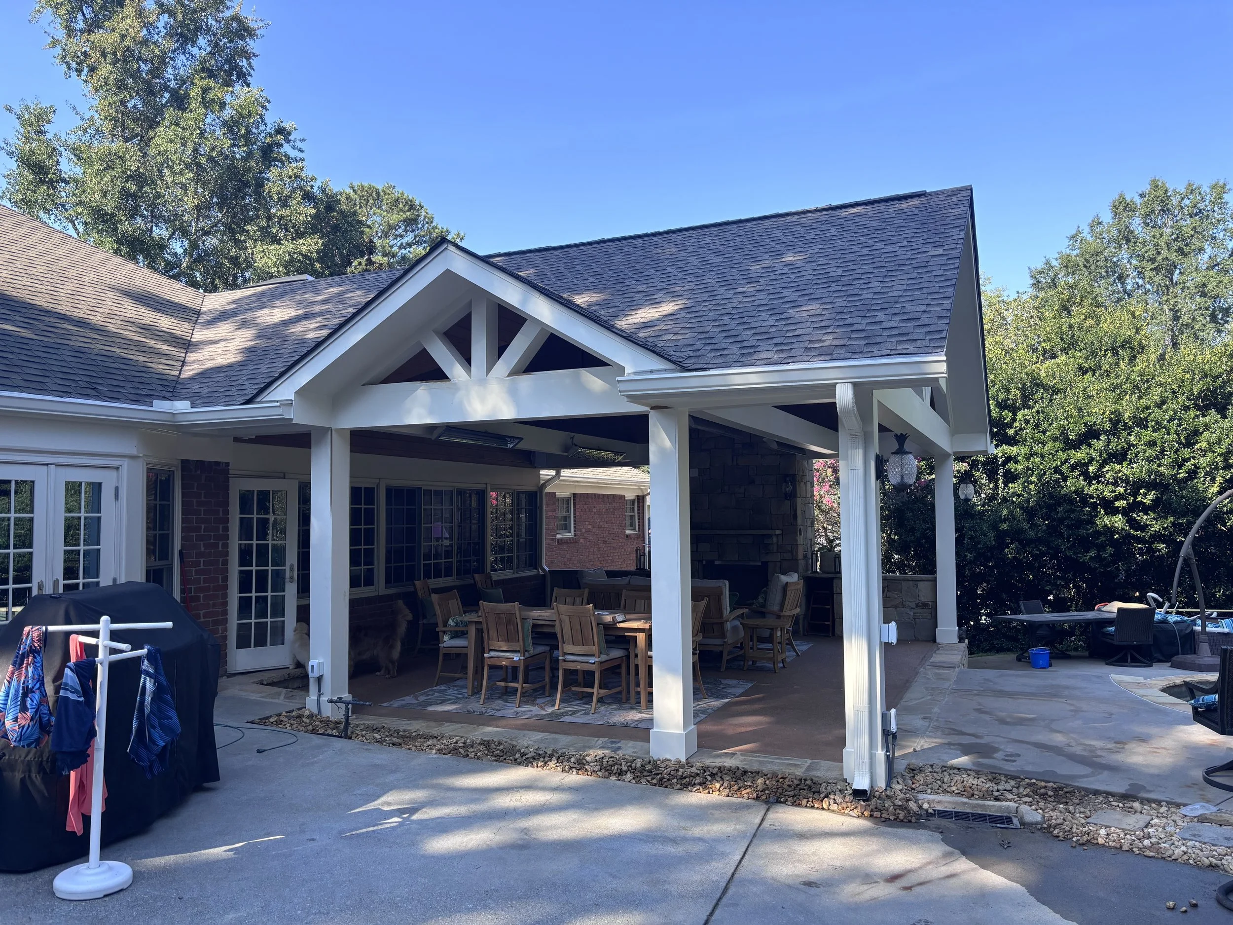 Backyard patio area with a covered dining space, wooden dining table and chairs, surrounded by trees and a clear blue sky.