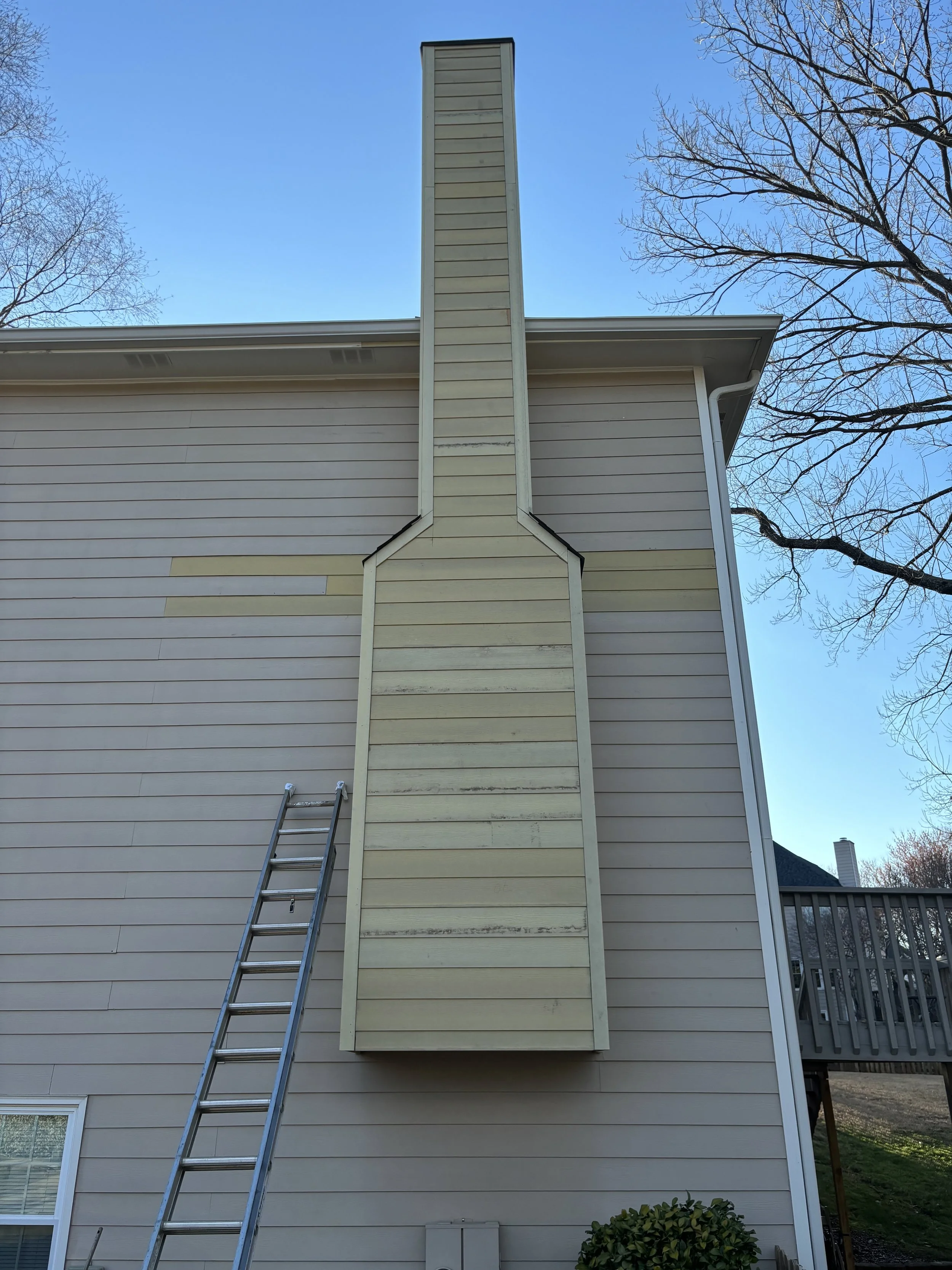 The image depicts the exterior of a house with beige siding, showing a tall, narrow chimney extending upward from the side of the house. A metal ladder leans against the house, reaching the chimney. The sky is clear and blue, with some trees visible on either side of the house and a small shrub at the bottom right corner.