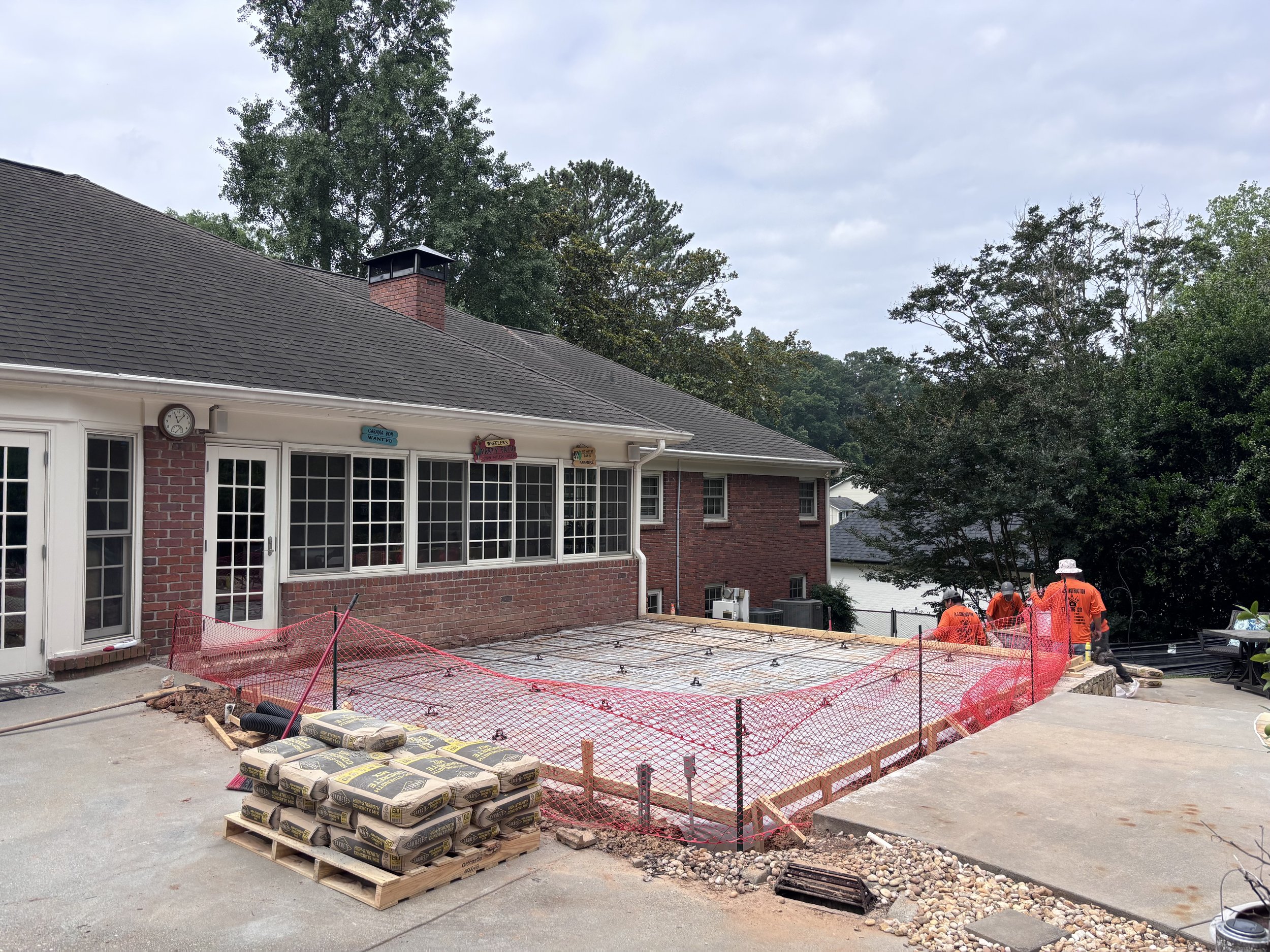 A house with a brick and white siding exterior, large windowed porch, and a chimney on the roof. The yard is under construction with orange safety fencing, scaffolding, and construction materials such as bags of concrete or mortar stacked on pallets.