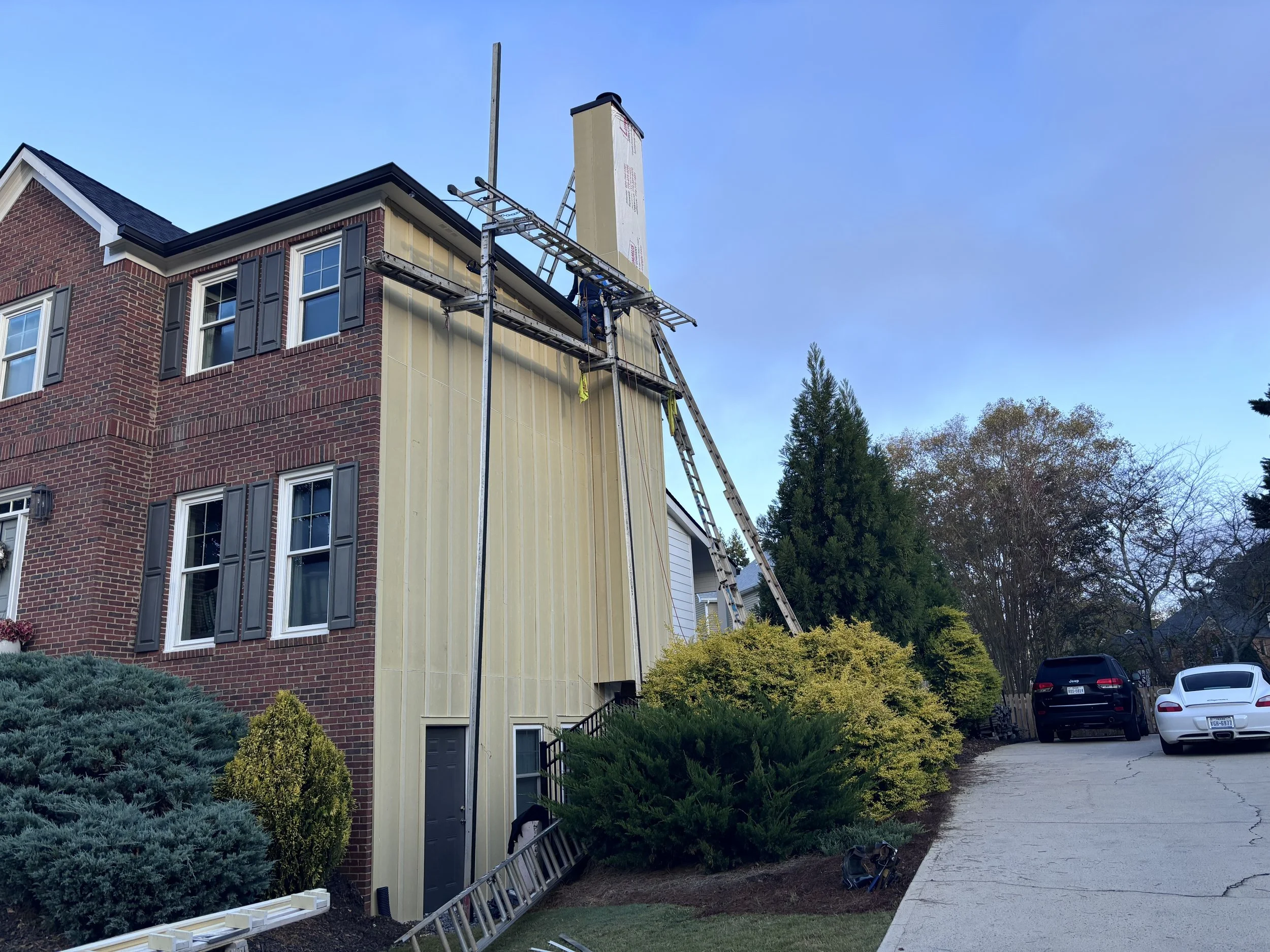 A house under renovation with workers installing siding on the upper part of the house. There are ladders leaning against the house, and the house has a brick and light-colored exterior. The yard has bushes and trees, and there are parked cars in the driveway.