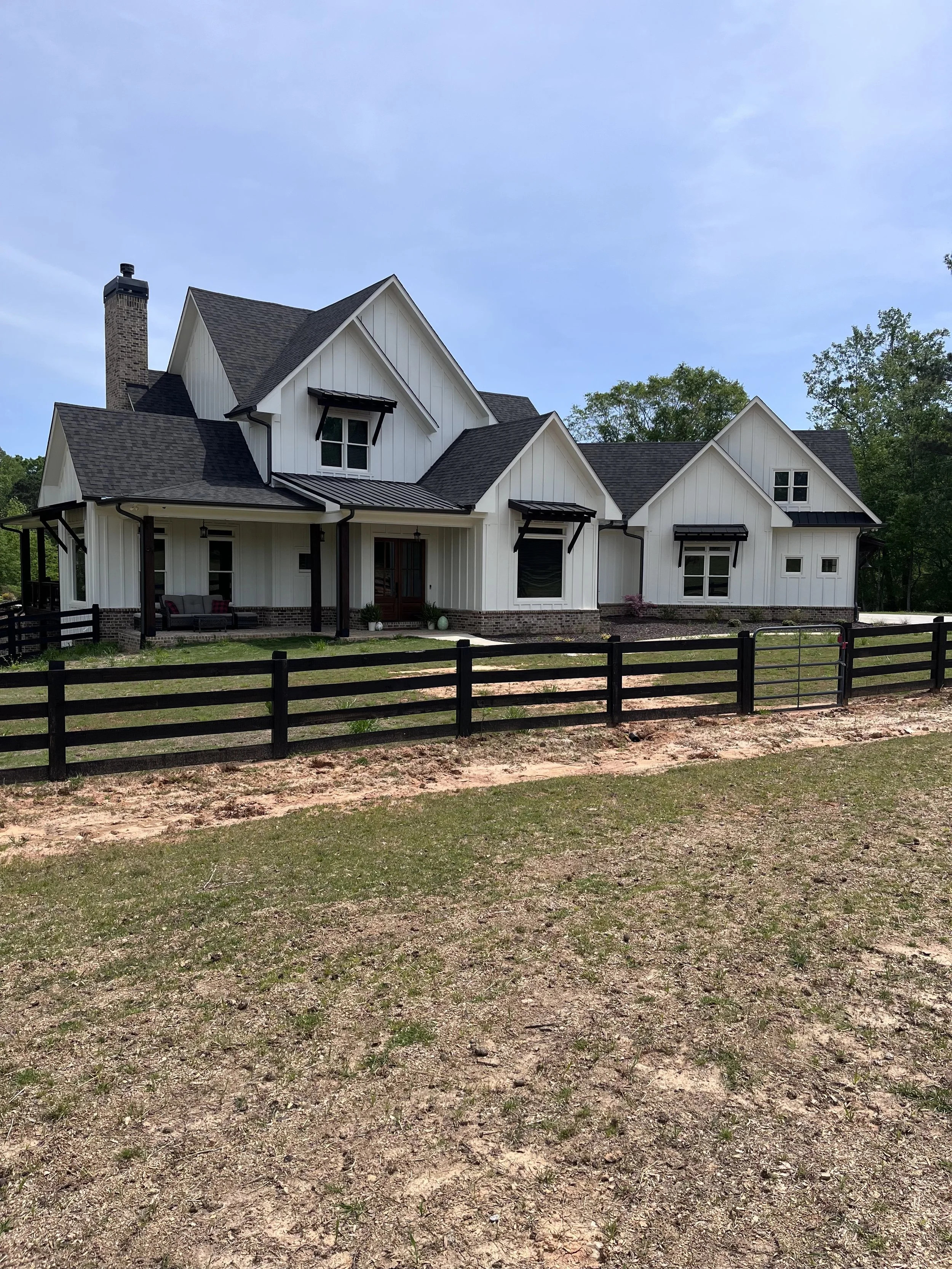 A modern white house with black accents, multiple gabled roofs, a chimney, and a front porch with seating, surrounded by grass and trees in the background.