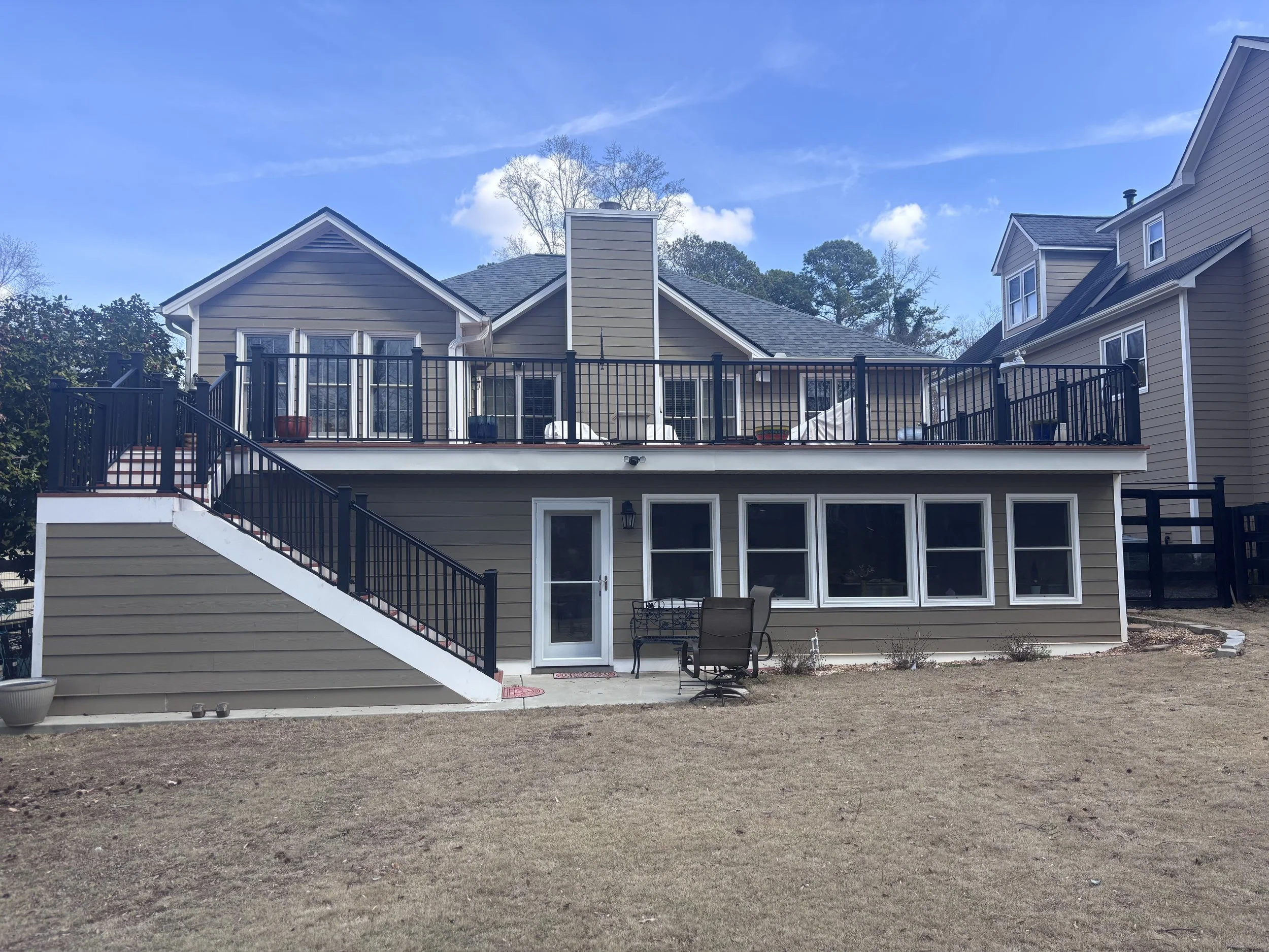 Backyard view of a beige house with a large deck, black railing, patio furniture, and sliding glass doors, under a blue sky with some clouds.