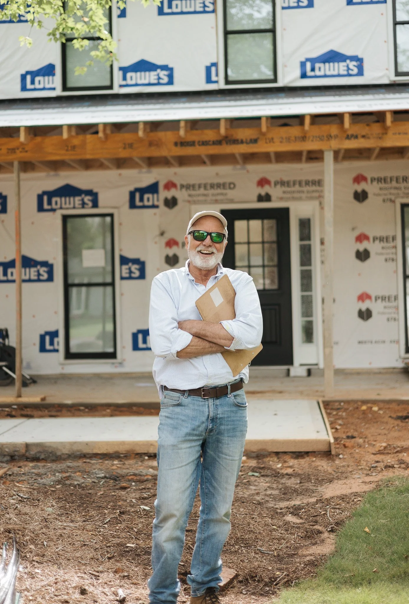 A man with a white beard, wearing sunglasses, a light blue shirt, and jeans, stands smiling with his arms crossed, holding a cardboard folder, in front of a house under construction wrapped with Lowe's and Preferred branding.