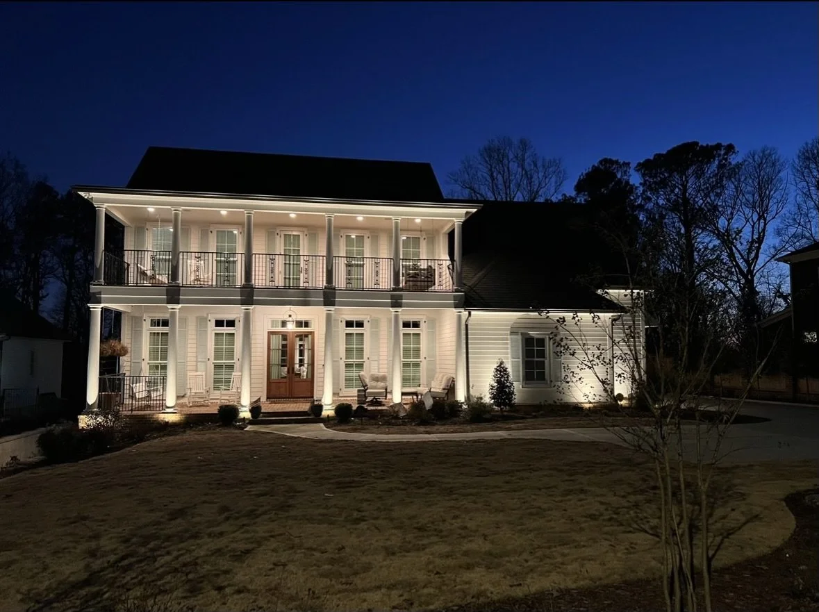 A well-lit two-story house at night with white siding, multiple windows with shutters, and a covered front porch with seating. The house is surrounded by a landscaped yard with small bushes and a tree, and the sky is dark blue.