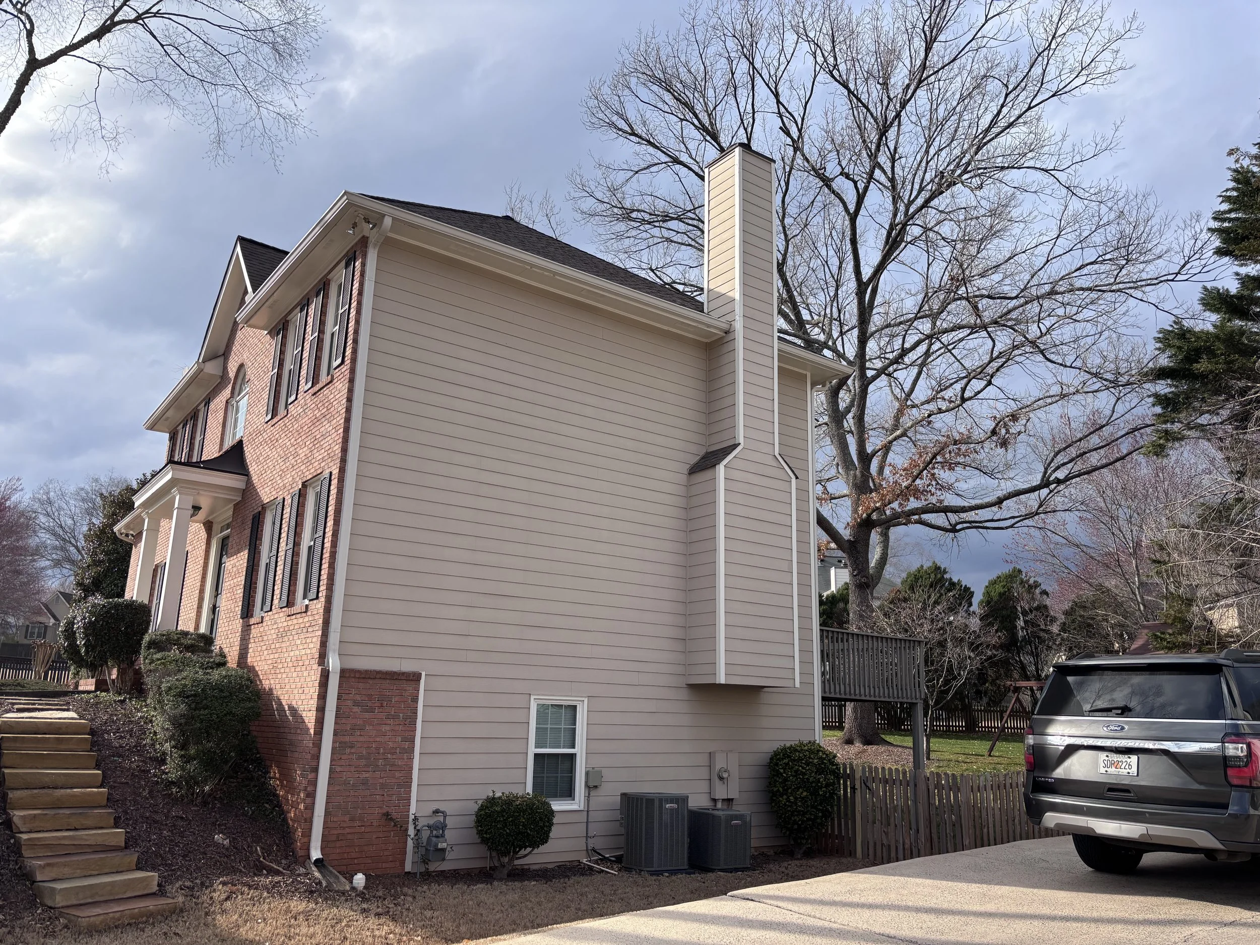The image shows the side view of a two-story house with a brick facade on the front and beige siding on the side. The house has dark shutters and a chimney. There are leafless trees in the background, a driveway with a parked SUV, and a small set of stairs leading to the front yard.
