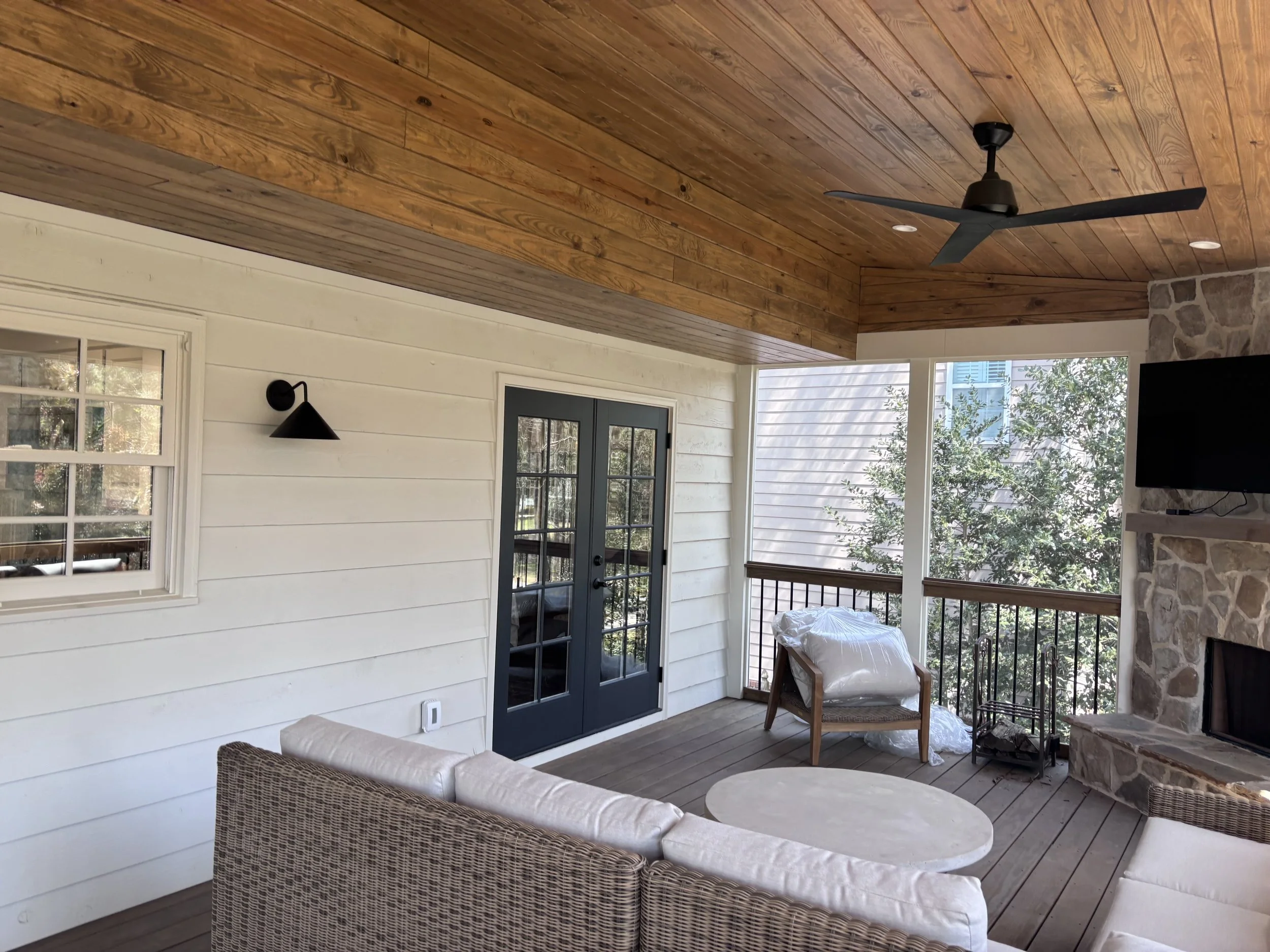 Covered porch area with white horizontal wooden siding interior walls, a stone fireplace, a ceiling fan, a black wall-mounted lamp, and doors and windows with black frames leading outside to trees and neighboring houses.