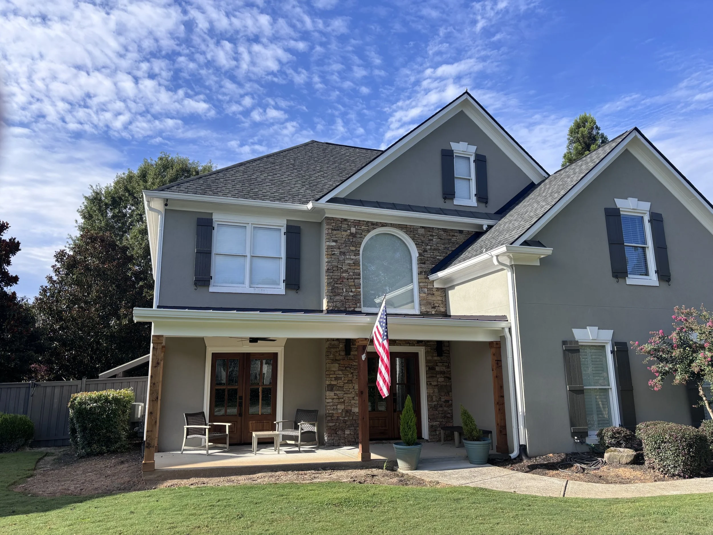 A modern two-story house with gray and stone exterior, large windows, a covered porch with outdoor seating, and American flag, set against a blue sky with clouds.