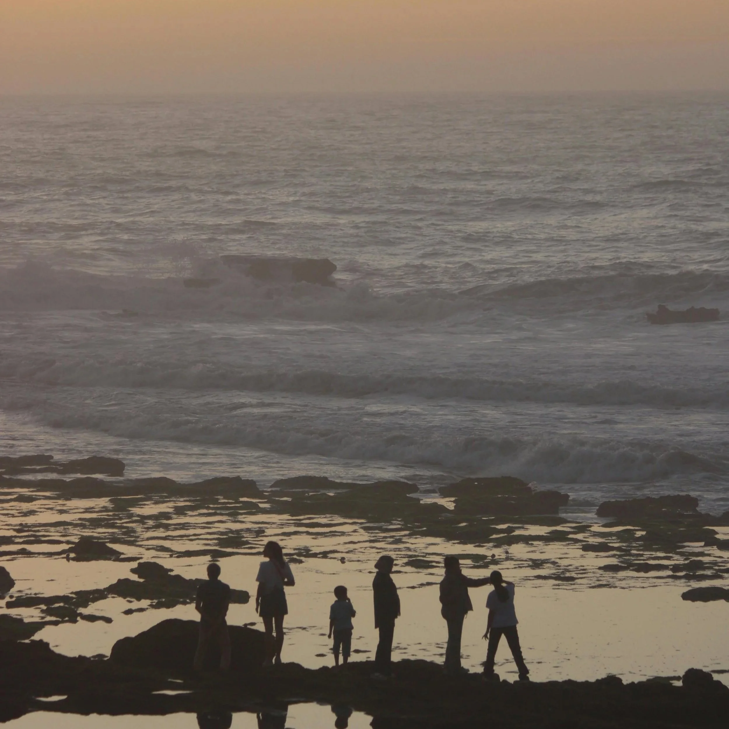 Silhouette of people walking along the rocky shoreline at sunset or dawn, with the ocean in the background and a hazy sky.