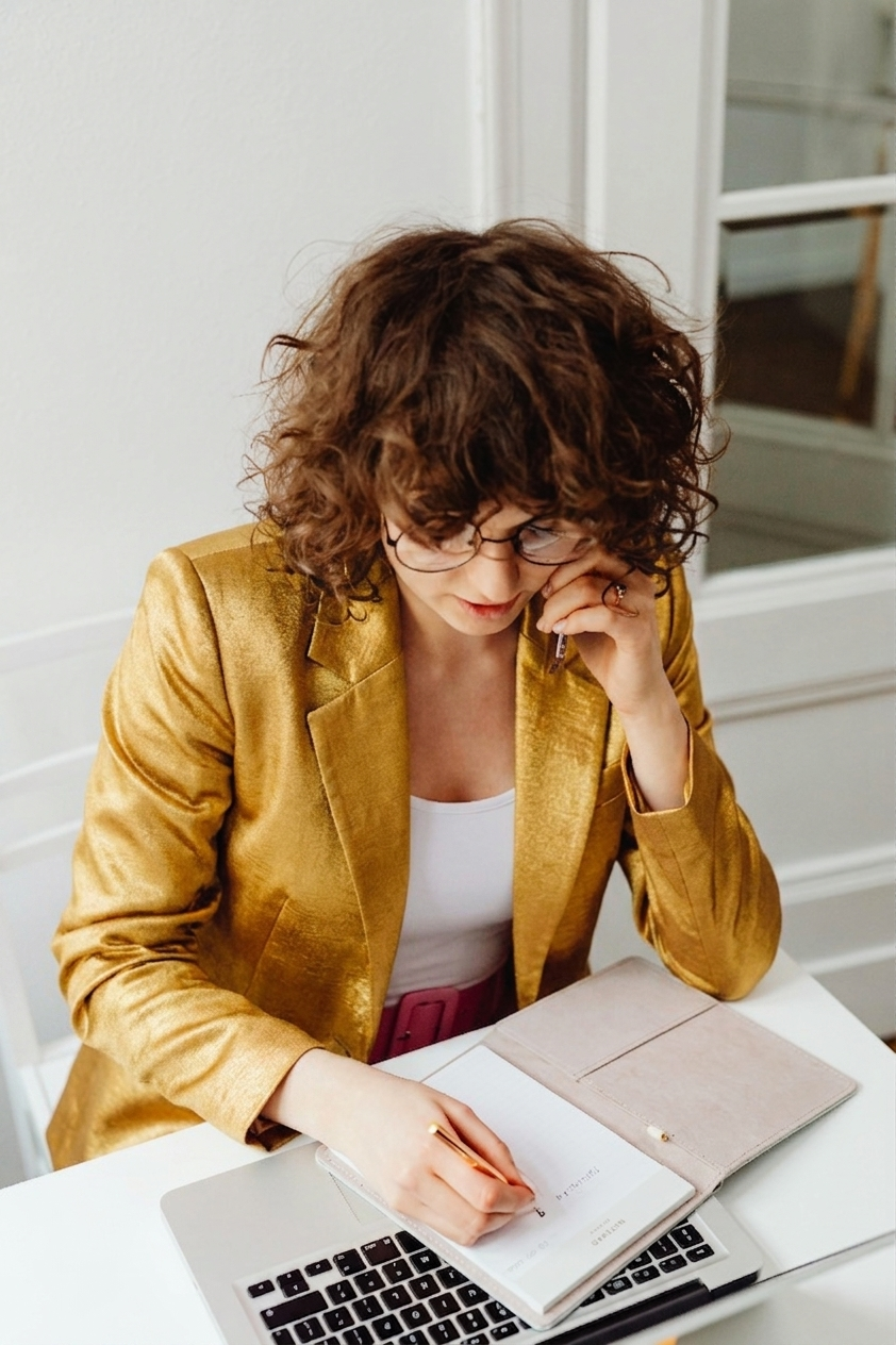 A woman with curly brown hair and glasses, wearing a gold blazer, sitting at a white desk, writing in a notebook with a pen, with a laptop and a beige clutch on the desk, in a home office setting.