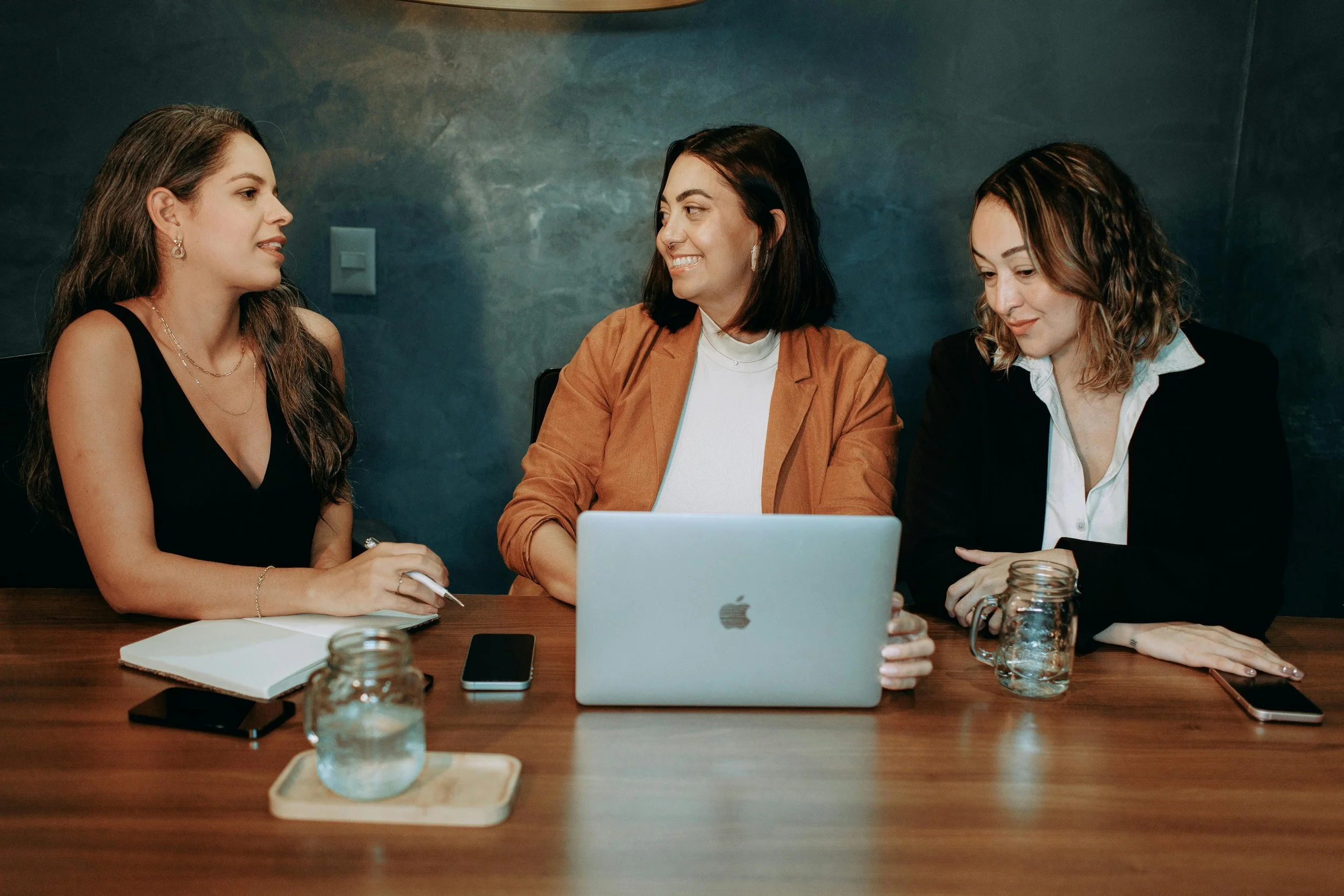 Three women sitting at a wooden table in a meeting room, engaged in conversation, with a silver laptop, notebooks, and smartphones on the table, and a dark painted wall in the background.