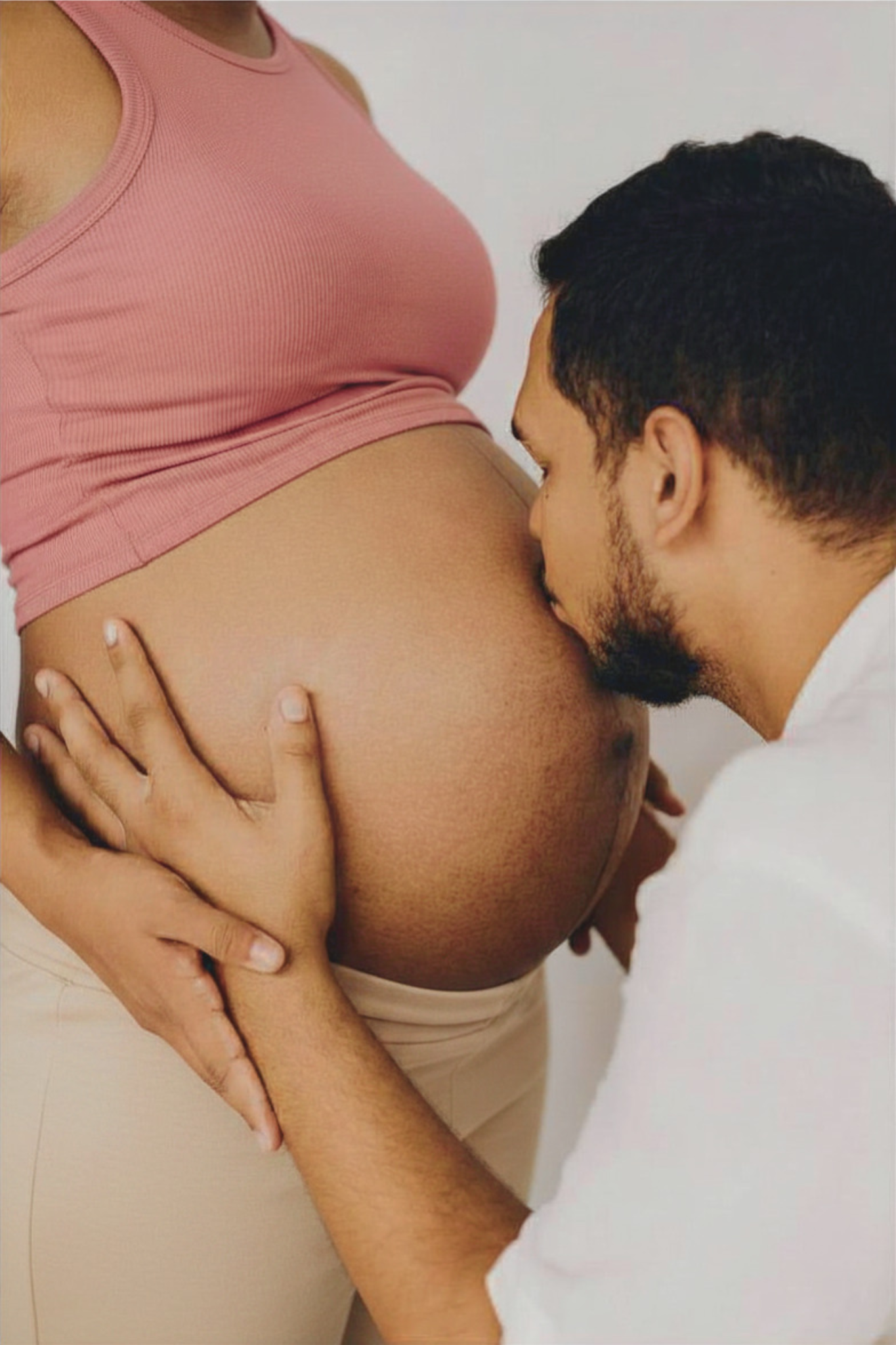 A person with dark hair and a beard is kissing the belly of a pregnant woman wearing a pink sleeveless top. The woman's hand is resting on her thigh.