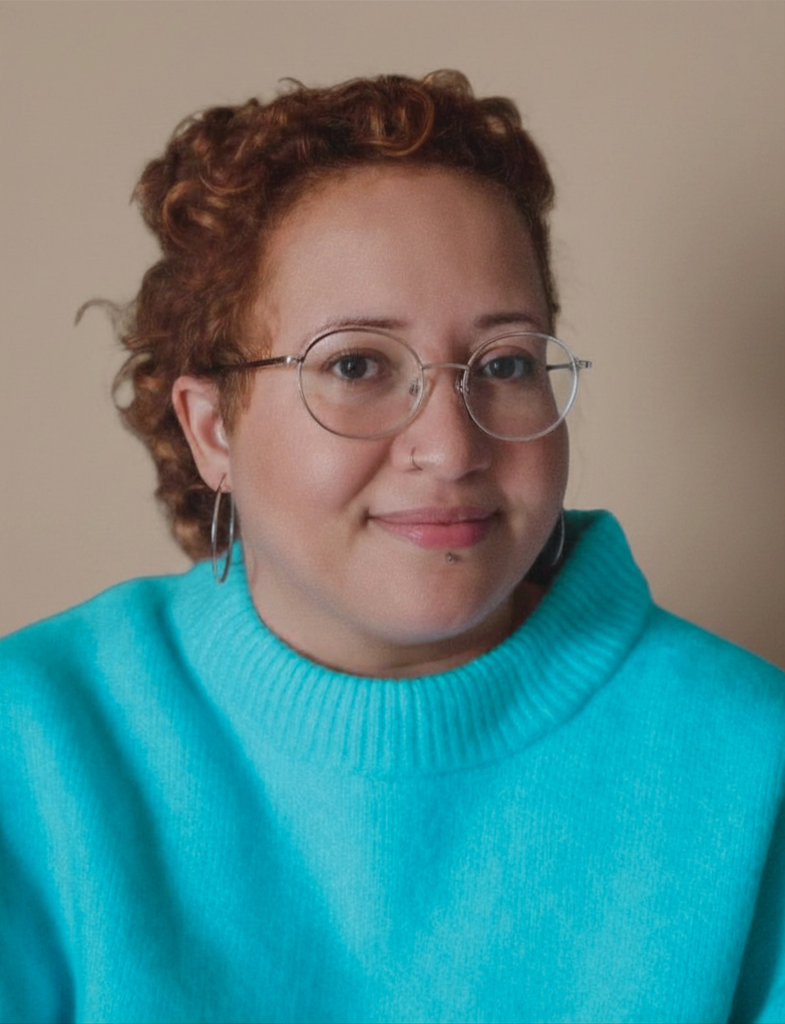 A person with curly red hair and round glasses smiles warmly at the camera, wearing a bright teal turtleneck jumper against a soft warm beige background.