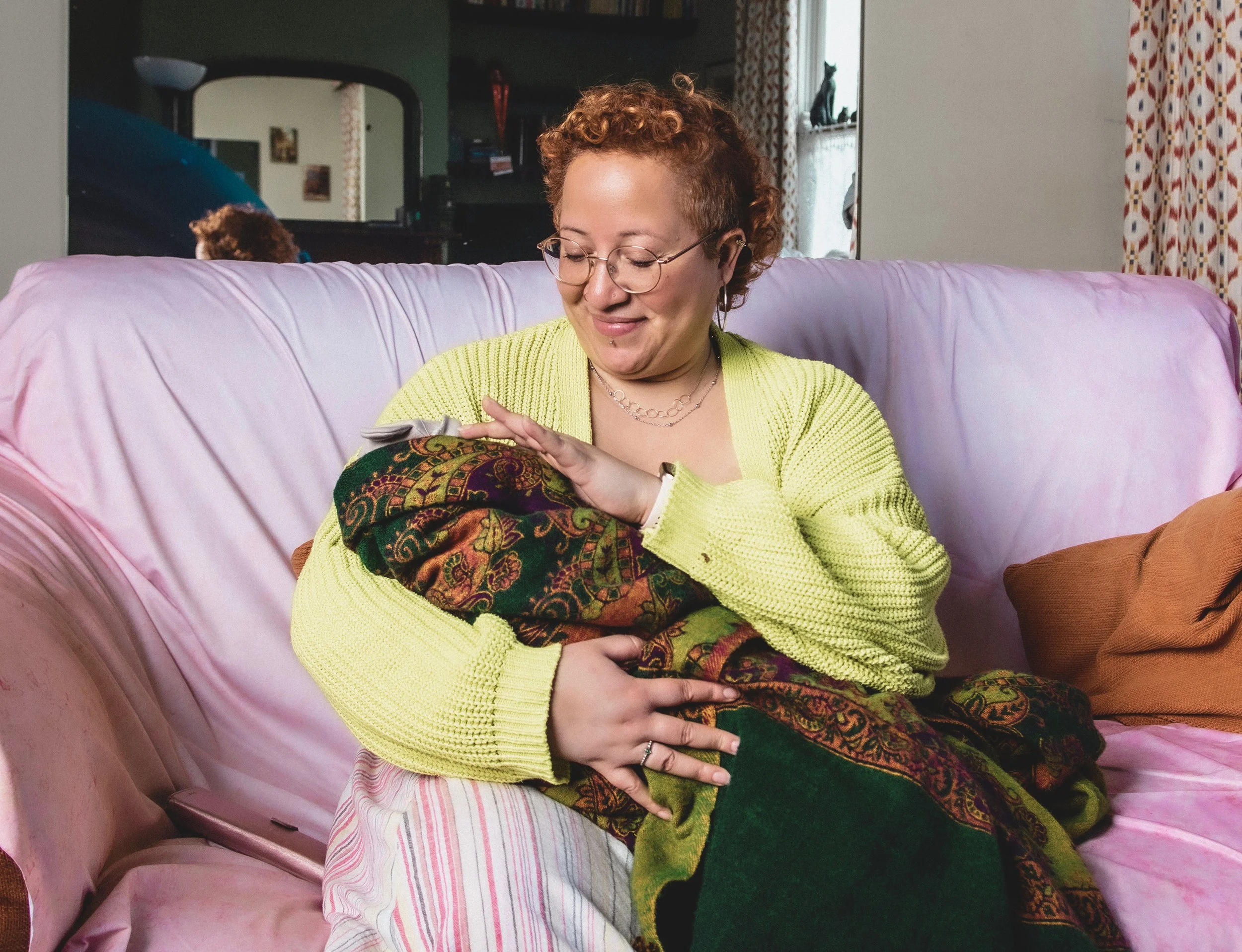 A person with curly red hair gazes tenderly at a newborn cradled in their arms, wrapped in a green and orange patterned shawl.
