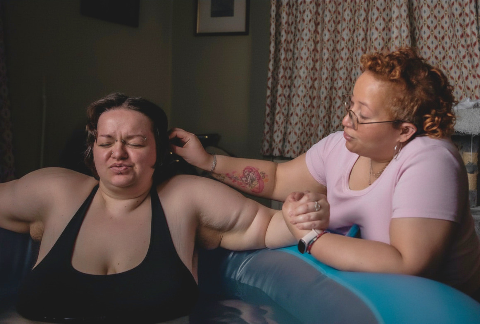 A person in a birthing pool concentrates through a contraction while a doula beside them holds their hand and gently brushes hair from their face.