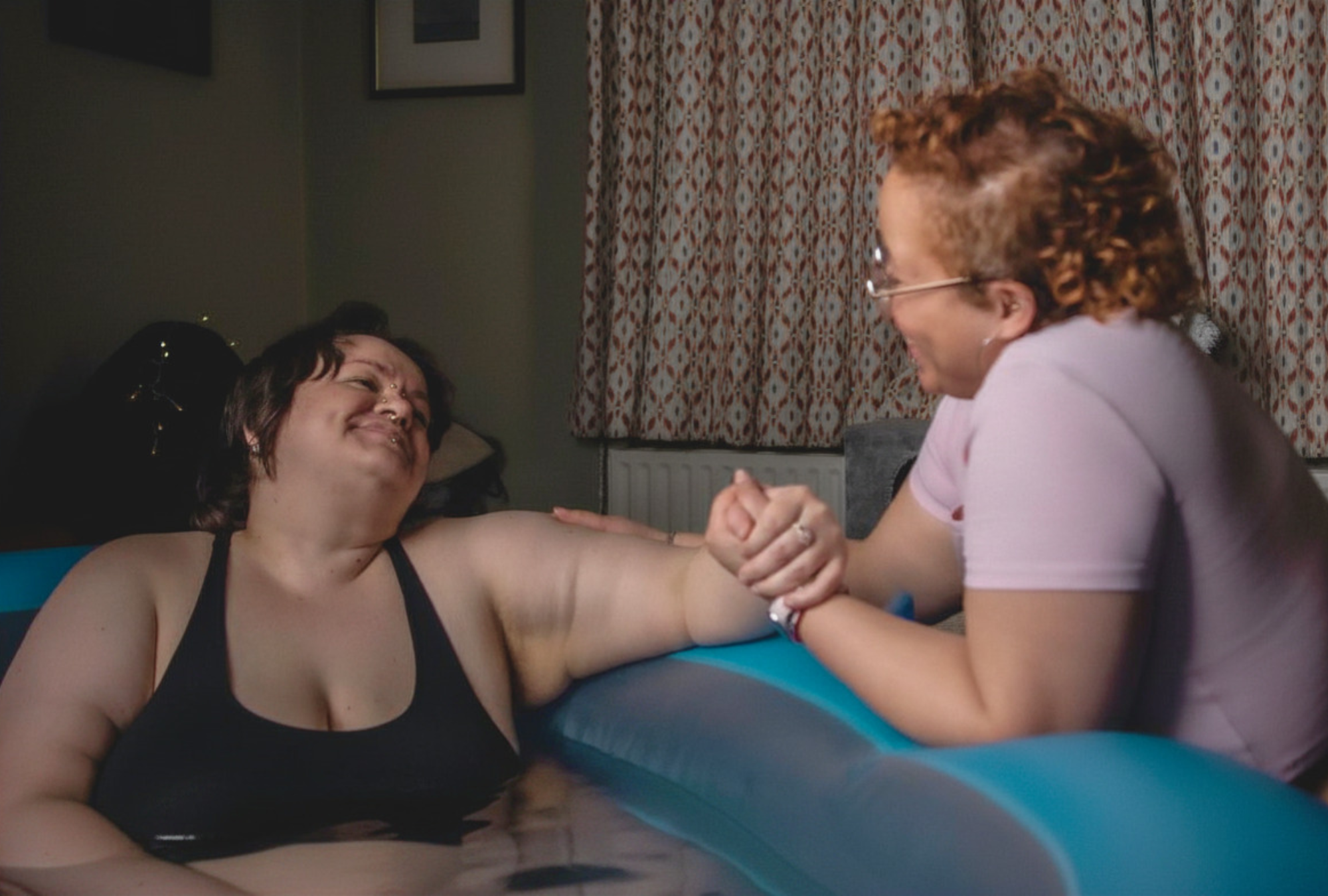 A person in a birthing pool smiles and grips the hand of a doula kneeling beside them, both sharing a warm moment during a home water birth.