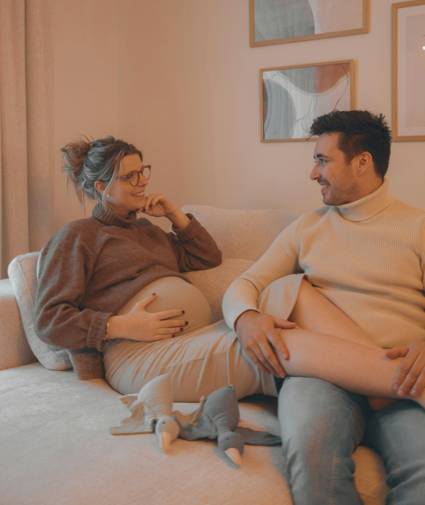 A pregnant woman with glasses and a brown sweater sitting on a couch, touching her belly and smiling, while a man in a cream turtleneck sweater sits beside her, smiling and looking at her, in a warmly decorated living room.