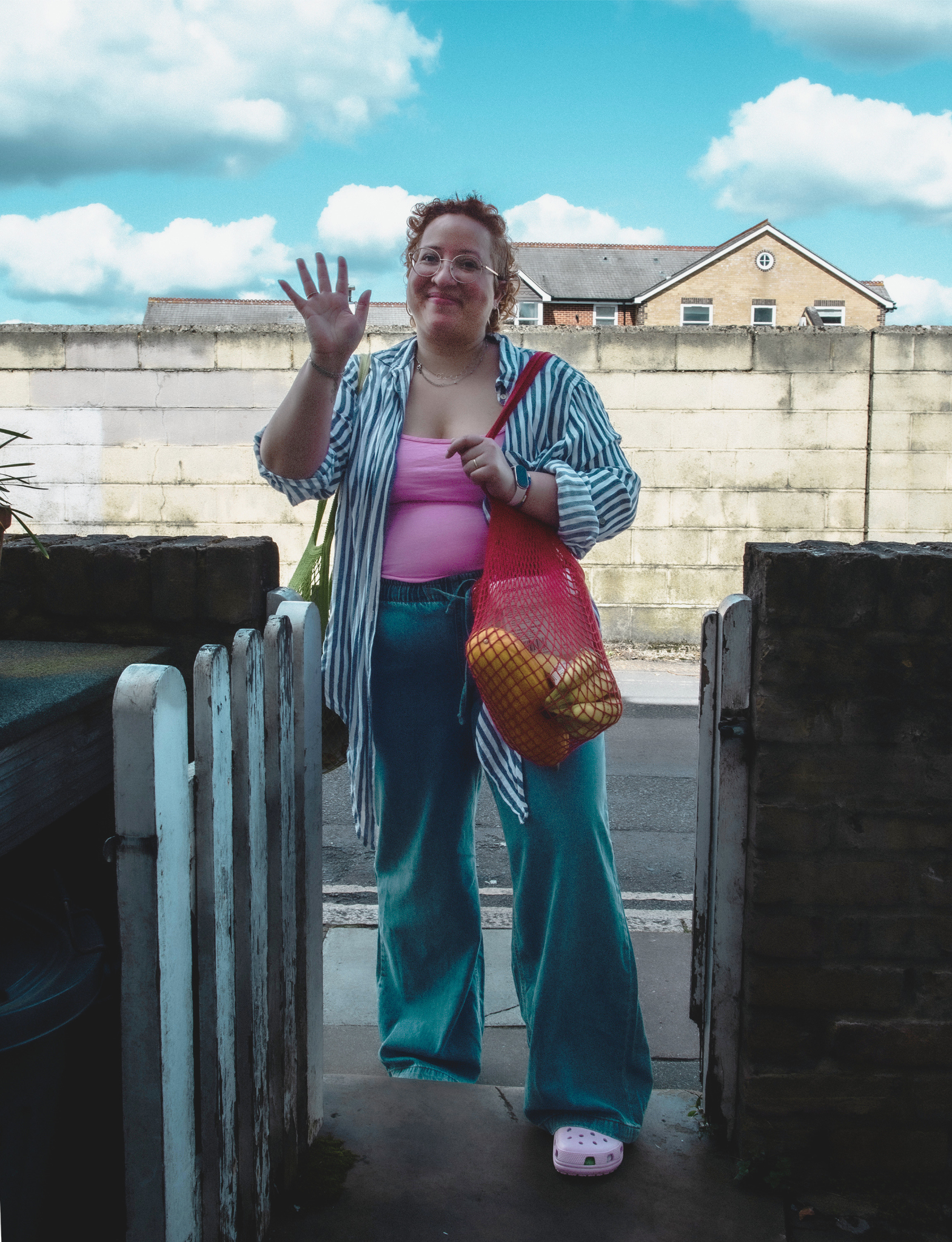 A person with curly red hair waves cheerfully at the door, carrying a red mesh bag of fruit, wearing a pink top, striped shirt, and wide-leg jeans.