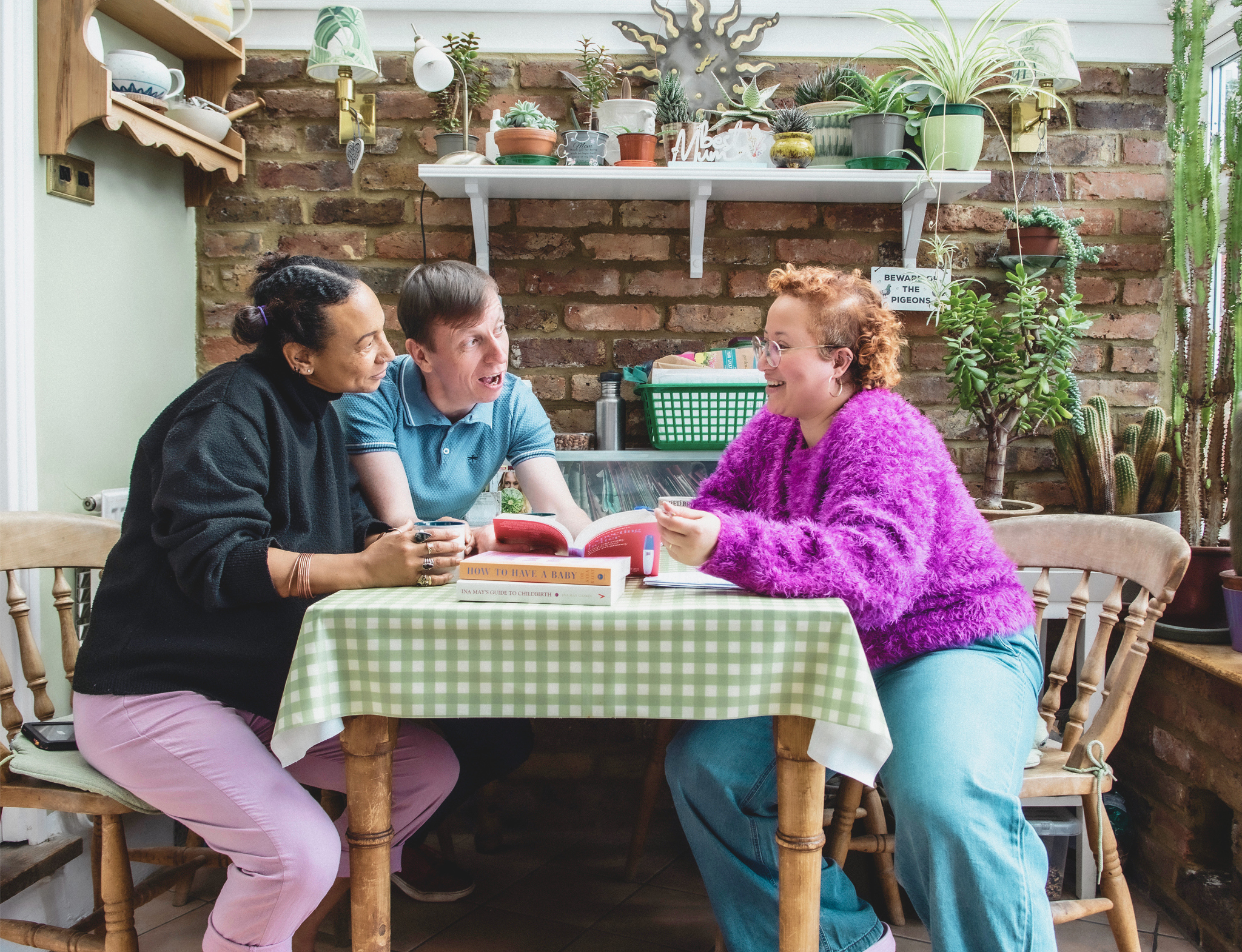 Three people laugh together around a table covered in birth prep books, in a cosy plant-filled kitchen with exposed brick walls.