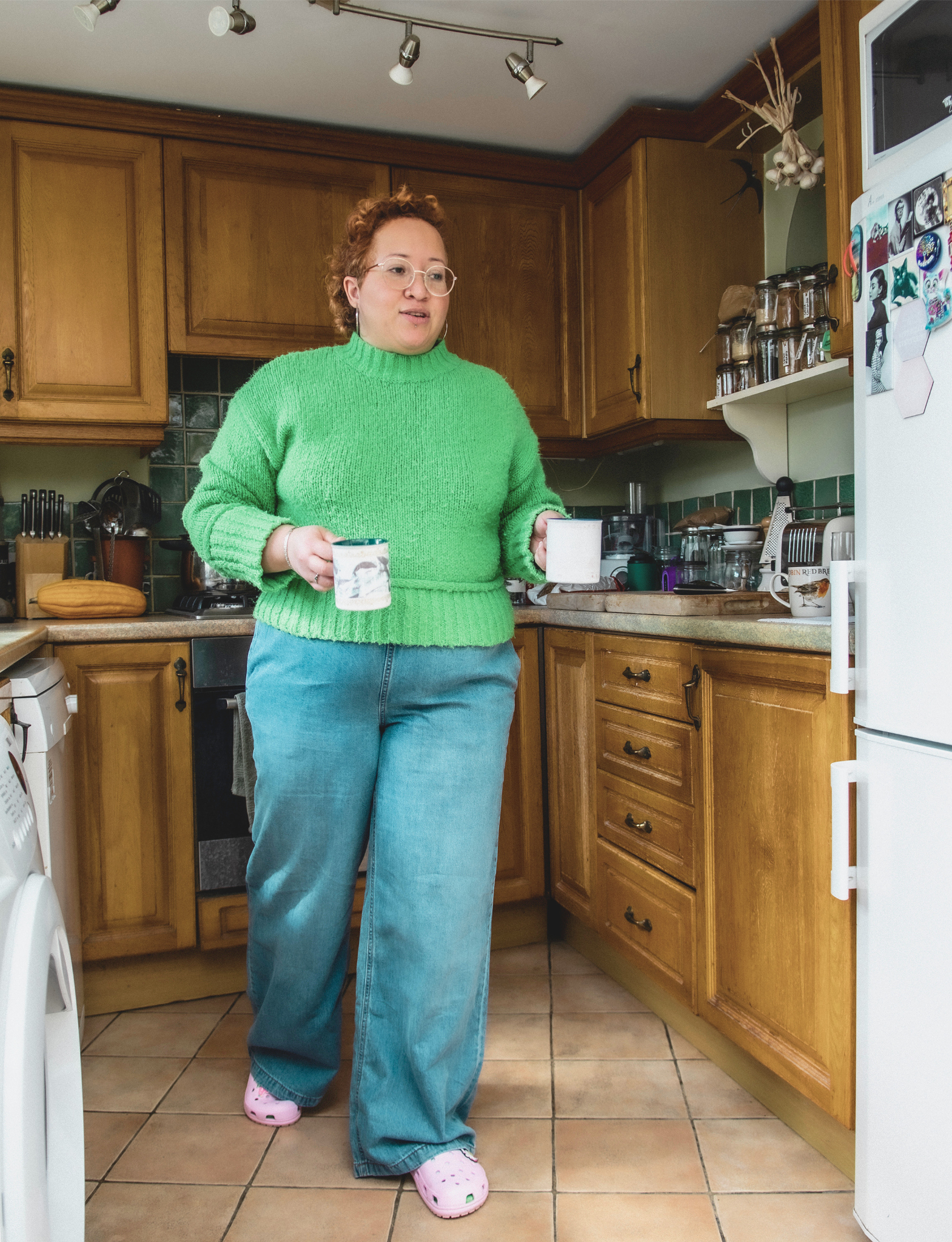 A person with curly red hair stands in a homely kitchen holding a mug, wearing a bright green knitted jumper, wide-leg jeans, and pink Crocs.