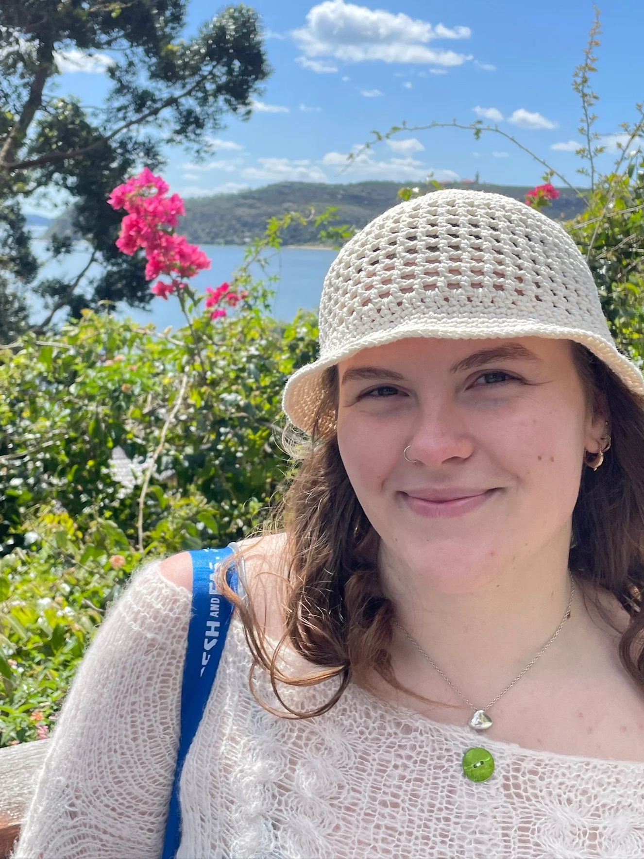 A woman smiling outdoors with the ocean, trees, and mountains in the background. She is wearing a white knitted top, a beige hat, and jewelry, and has a blue bag on her shoulder.
