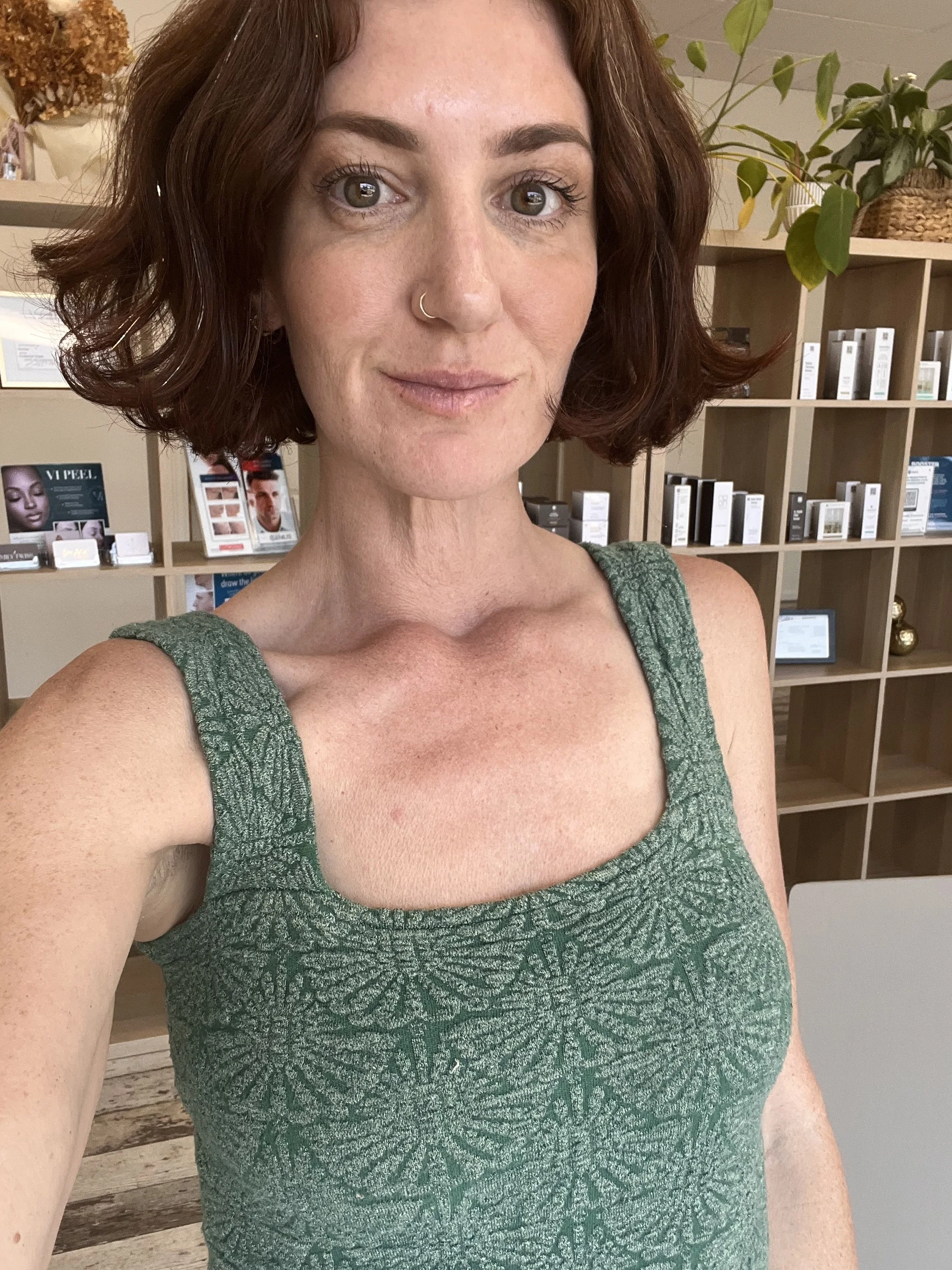 A woman with shoulder-length curly auburn hair, fair skin, and freckles taking a selfie indoors, with wooden shelves and plants in the background.