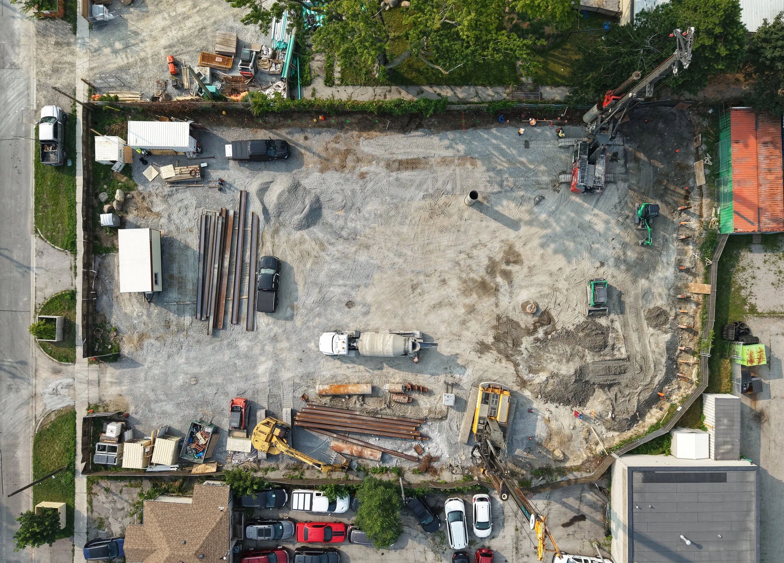 An aerial view of a construction site with various construction vehicles, equipment, and materials, surrounded by residential houses, trees, and parked cars.