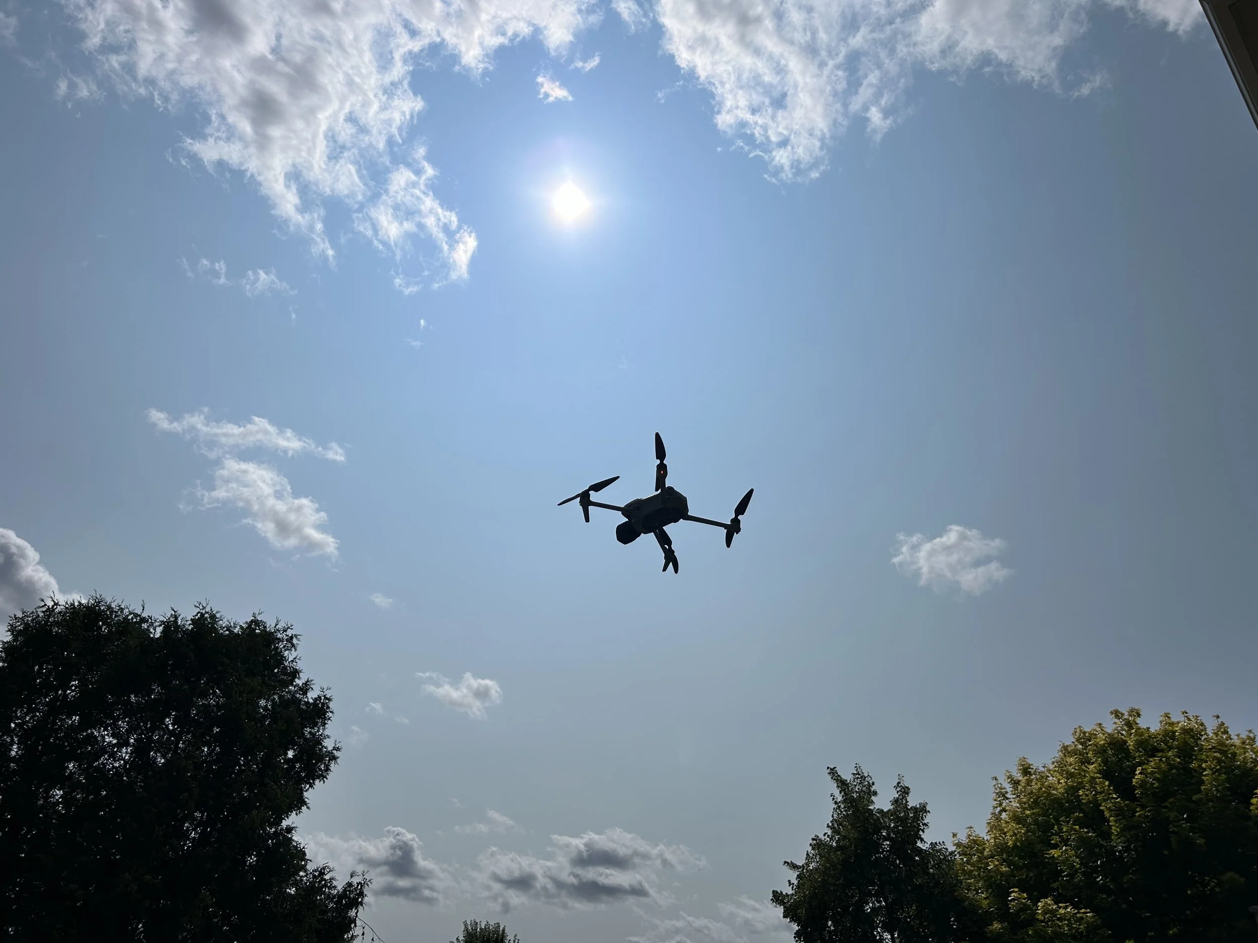 A drone flying in the sky with clouds and the sun visible, trees at the bottom of the image.