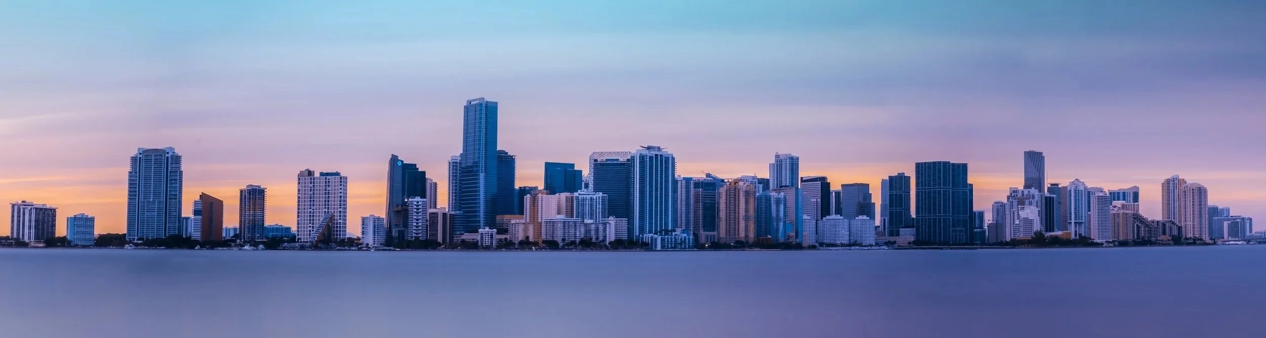 A city skyline with tall modern skyscrapers at sunset, viewed from across a body of water.