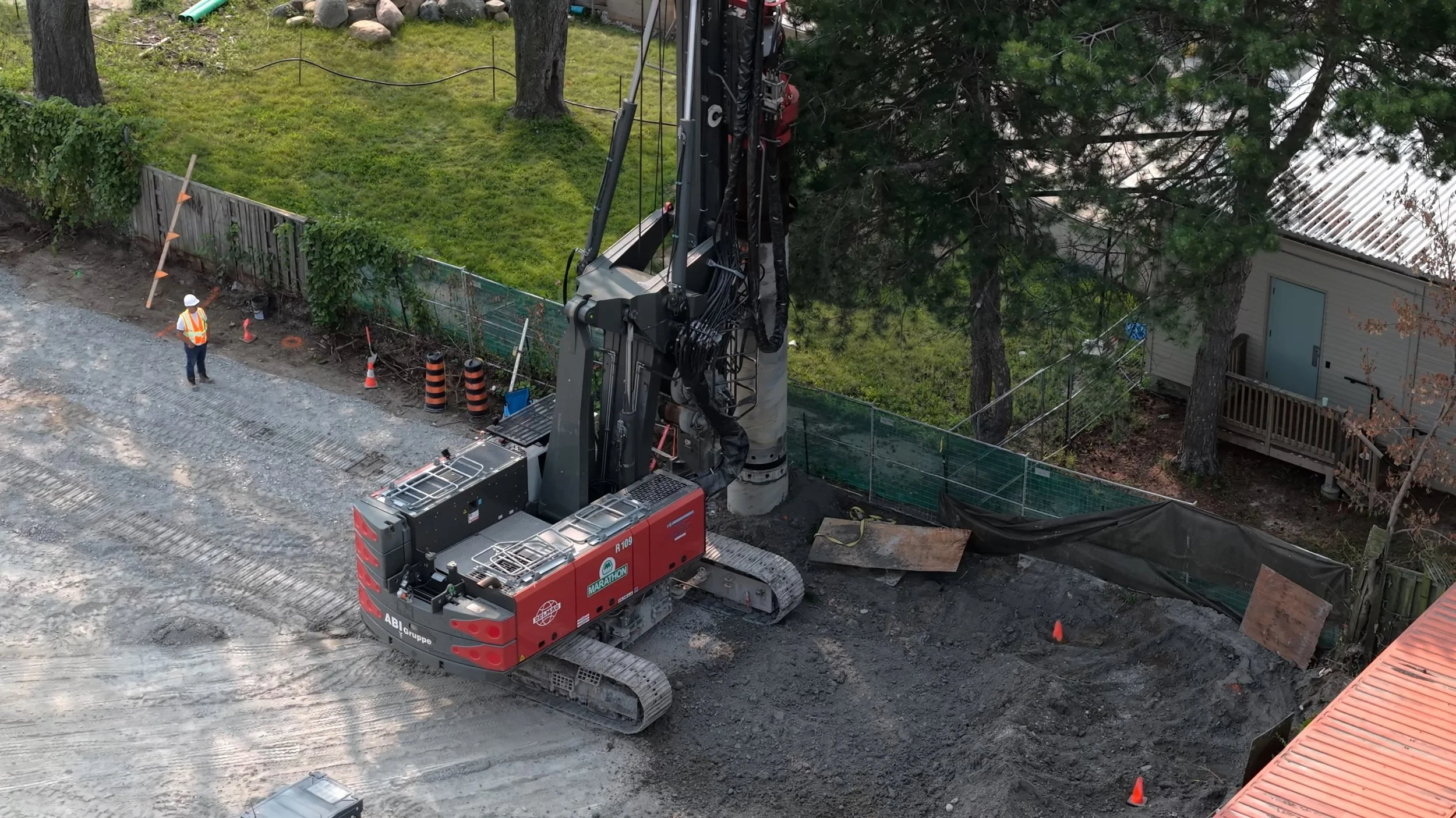 An underground drilling machine at a construction site, with a worker in a safety vest and helmet nearby, dirt ground, and a green backyard with trees and a house in the background.