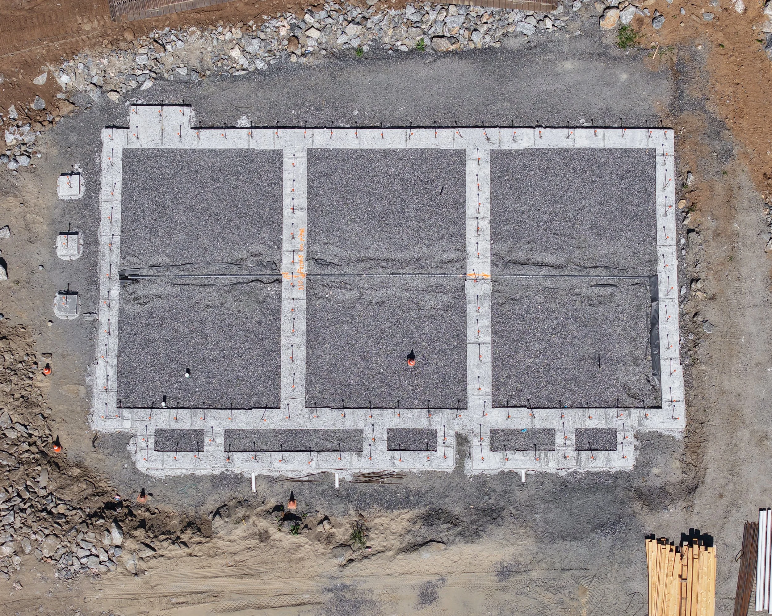 Aerial view of a construction site with a foundation outline marked in concrete and gravel, surrounding dirt and rocks, and nearby construction materials and equipment.