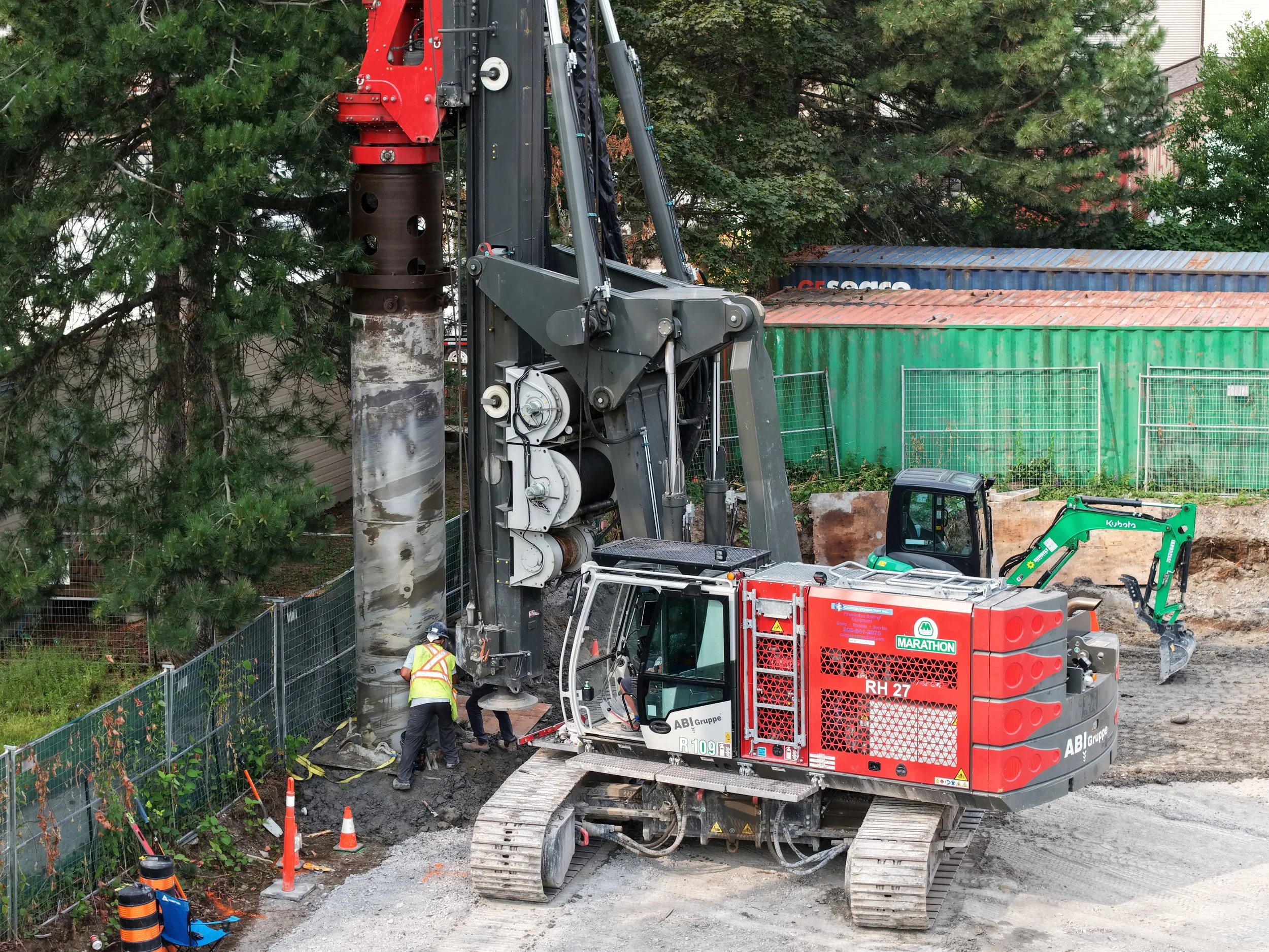 A construction site where a large drilling machine is working on a concrete column surrounded by trees and a green fence. Workers in safety vests and helmets are present, and there are various construction tools and materials nearby.