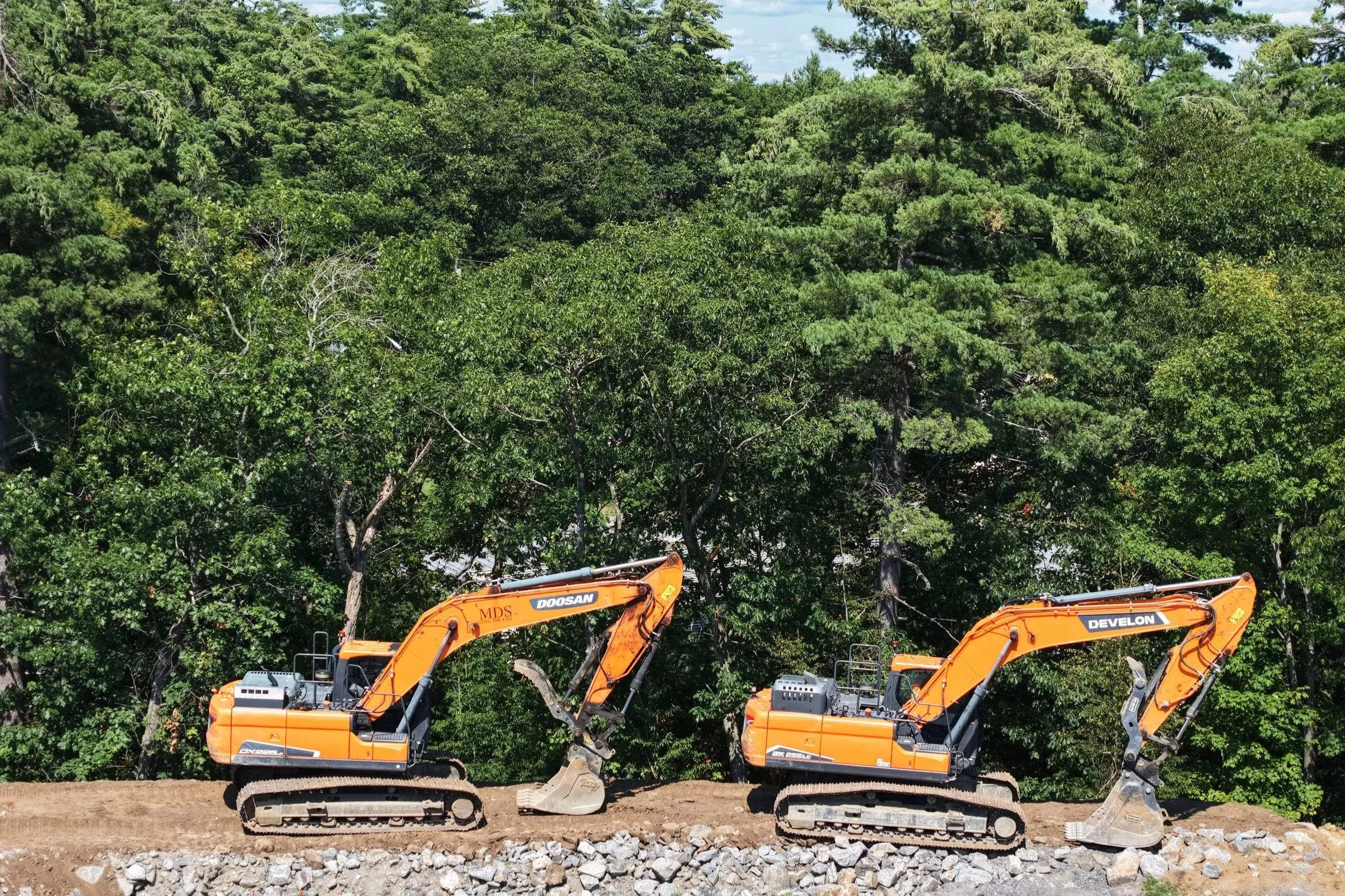 Two orange construction excavators working on a construction site with a dirt ground and large rocks in front, set against a background of dense green trees and a partly cloudy sky.