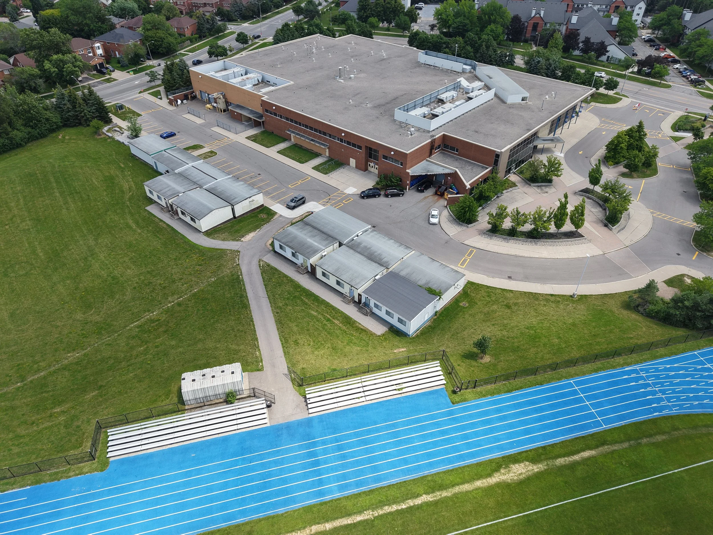 An aerial view of a school or stadium with a blue running track, white benches, a grassy field, a brick building with a flat roof, parking lot, and surrounding residential neighborhood with trees.