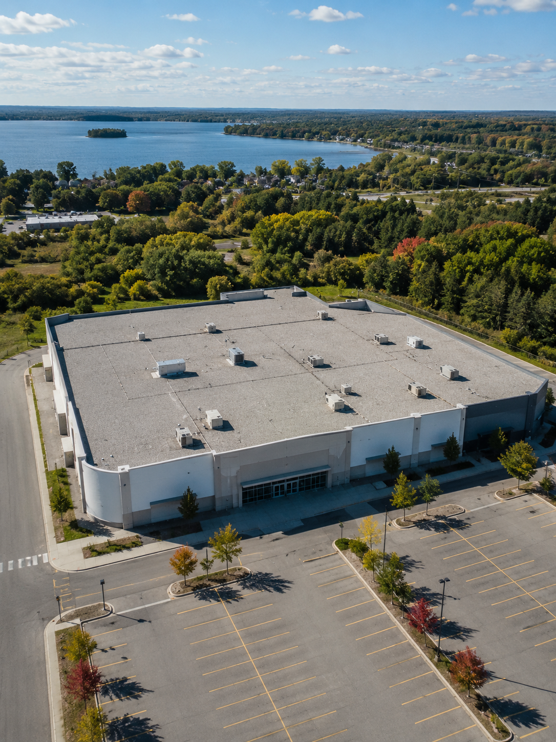A large, empty parking lot in front of a commercial building with a flat roof, surrounded by trees, with a lake and a forested landscape in the background under a partly cloudy sky.