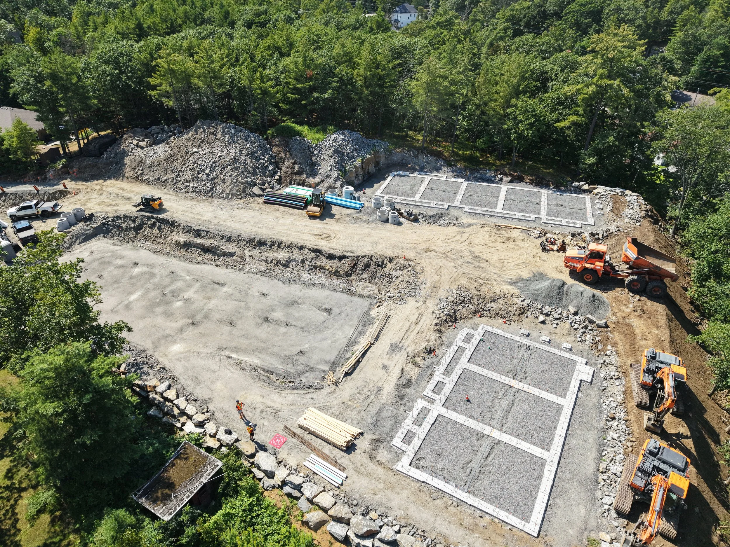 Aerial view of a construction site with foundations being poured, surrounded by trees, construction vehicles, and building materials.