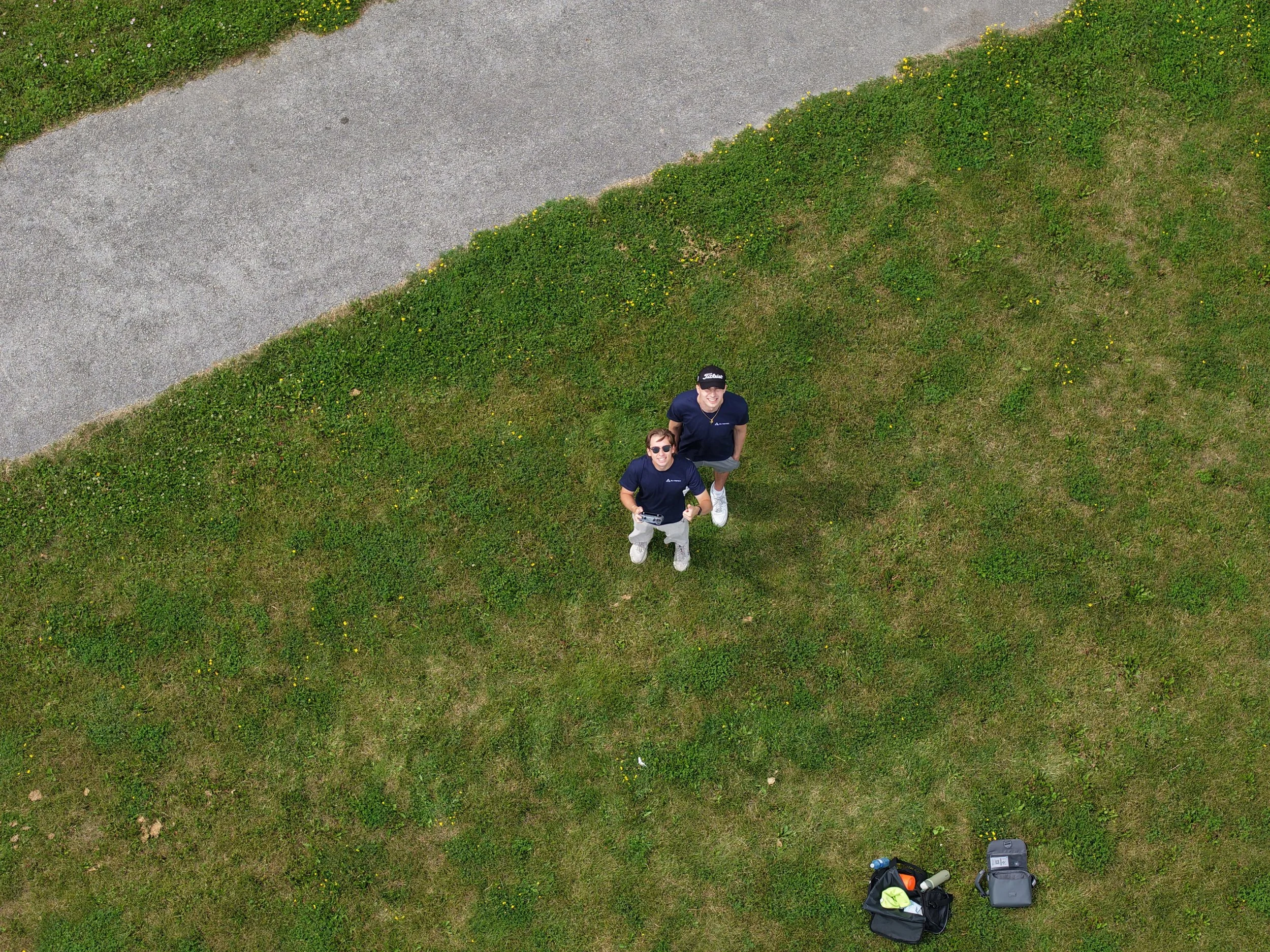Two people standing outdoors on a grassy area with small yellow flowers, looking up at the camera. There are bags and bottled drinks on the ground nearby.