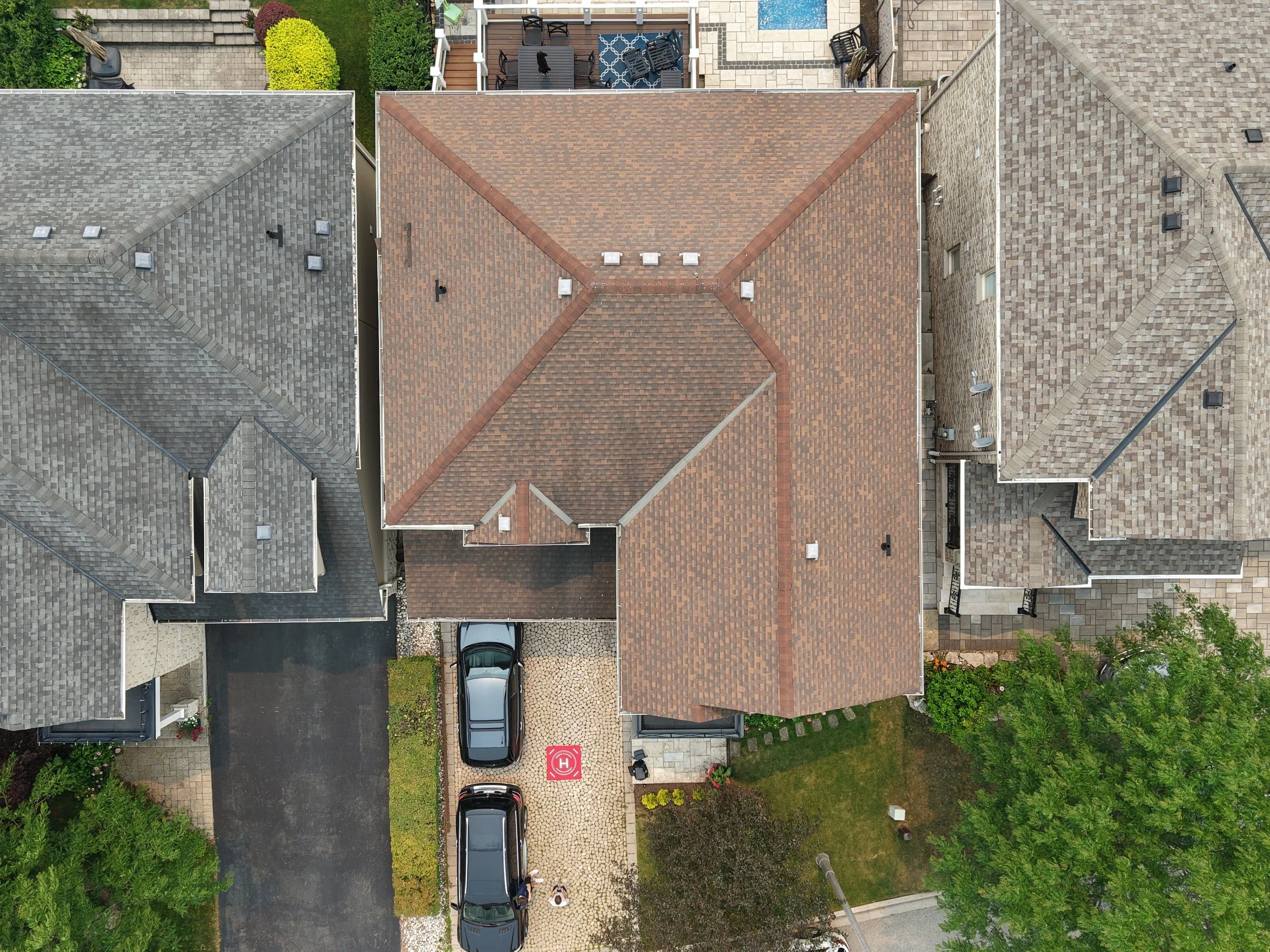 Bird's eye view of rooftops and backyard patios of multiple houses, with parking area, greenery, and a red helicopter landing pad.