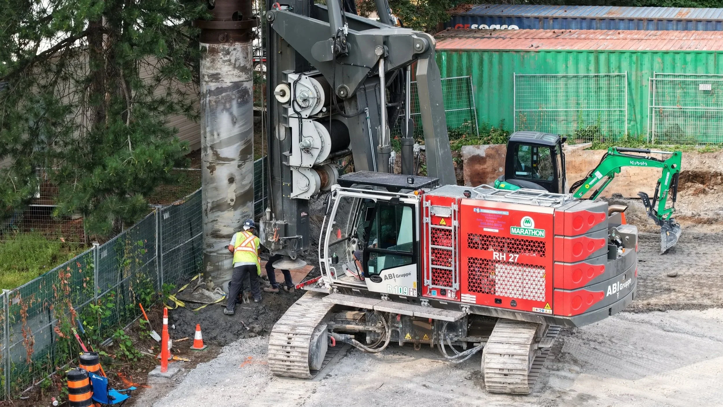 Construction workers and machinery at a site, with a large drilling machine and a mini excavator working near a fence and trees.