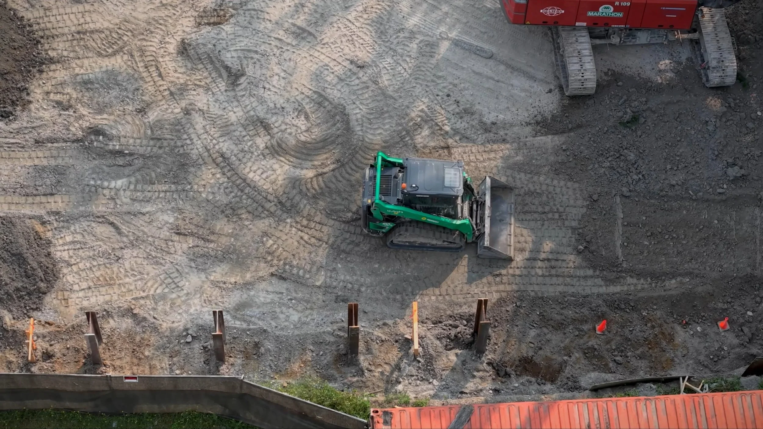 An overhead view of a construction site with a small green and black excavator working on the dirt, leaving tracks and patterns. The site is bordered by a low wooden barrier with orange cones and posts, and there's a section of bright orange safety fencing at the bottom.