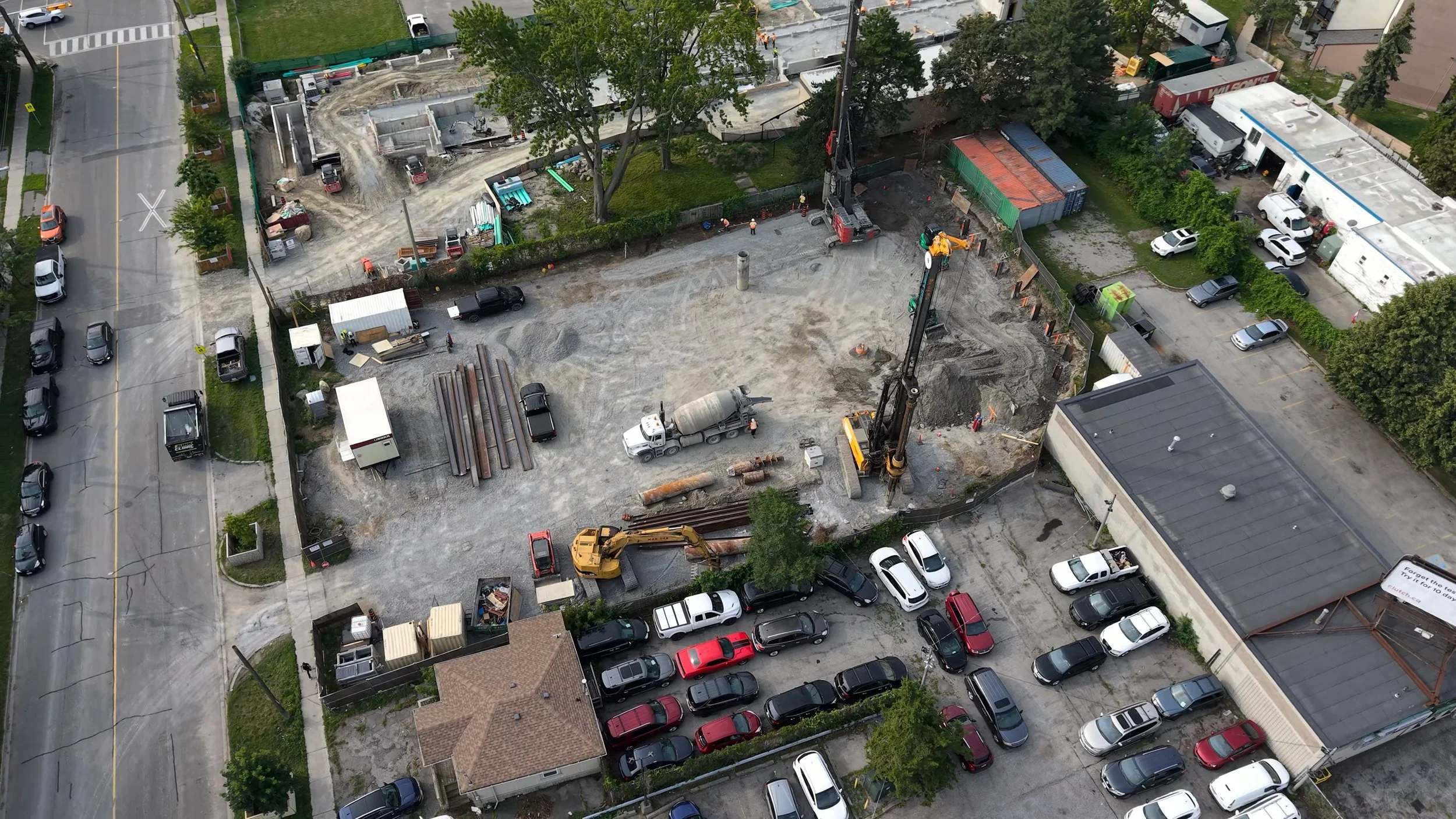 Aerial view of a construction site with heavy machinery, construction workers, and surrounding parked cars, located between residential and commercial buildings.