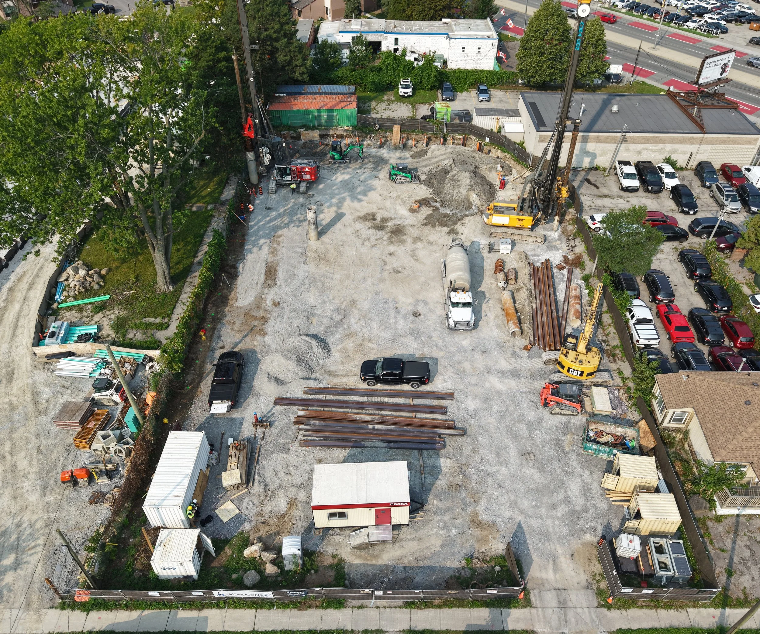 Construction site with large drilling machine and excavator, surrounded by parked cars, trees, and nearby buildings.