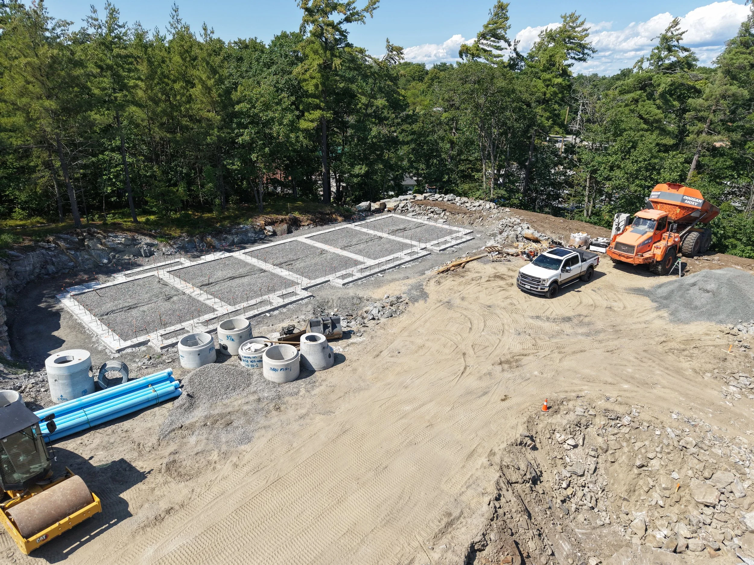Construction site with leveled ground, framing of foundation with wooden stakes, large orange cement mixer truck, white pickup trucks, pipes, concrete cylinders, and equipment, surrounded by trees and a partly cloudy sky.