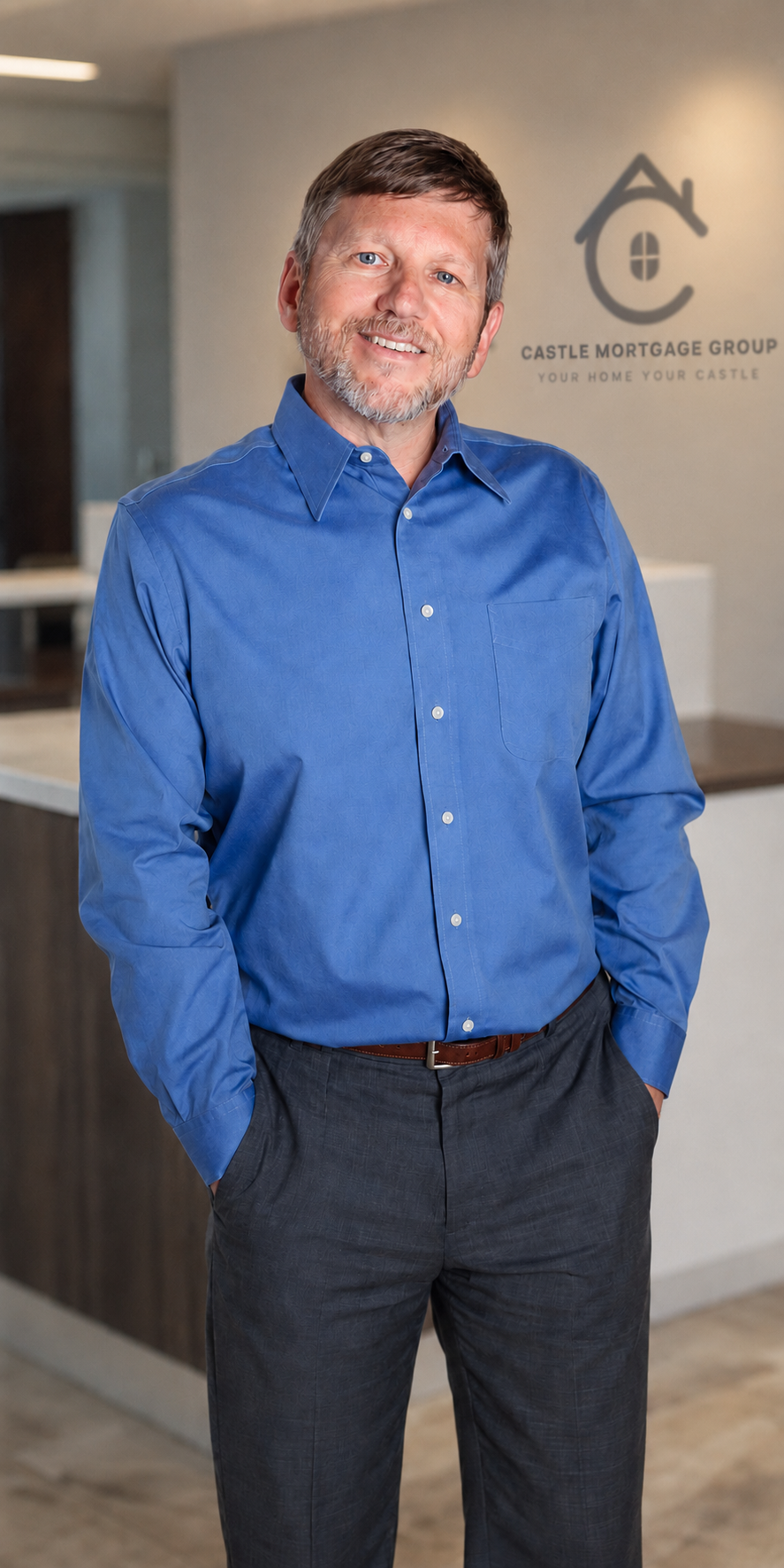 A man with gray hair and beard wearing a blue button-up shirt and dark trousers standing in front of a wall with the Castle Mortgage Group logo.