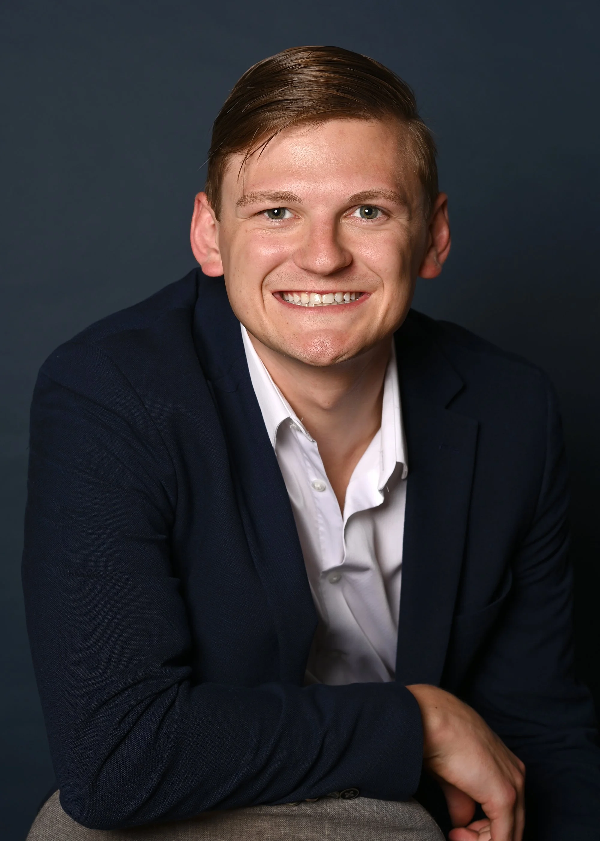 Portrait of Devin Carver, smiling and wearing a dark blazer and white shirt, sitting against a dark background.