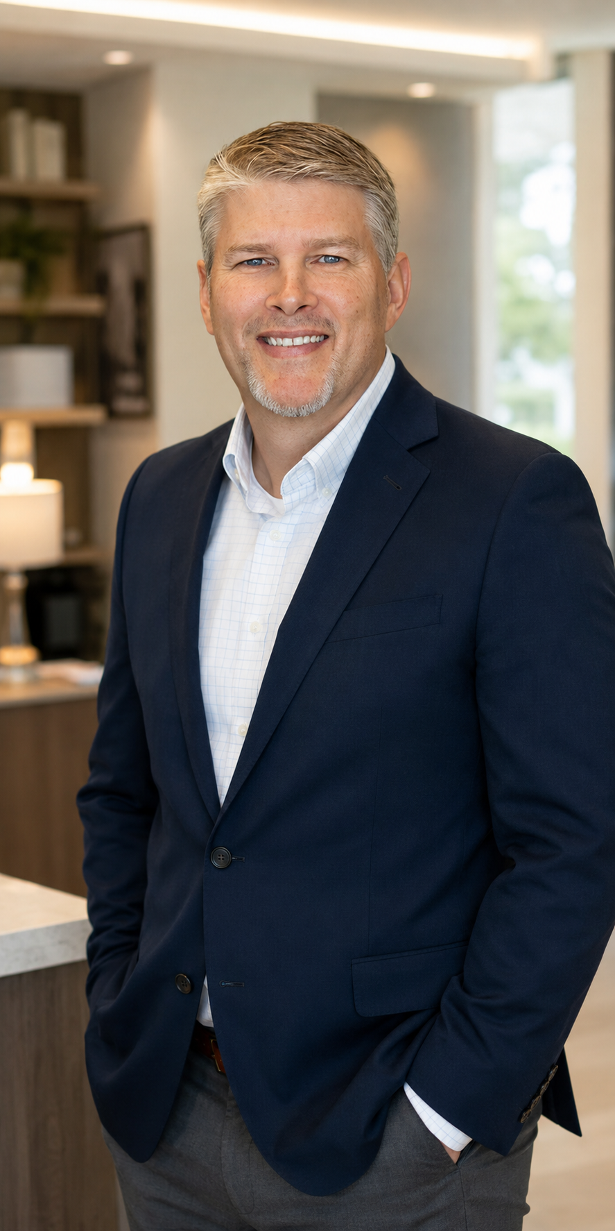 Professional man with gray hair and a goatee smiling in an office setting, wearing a navy blue suit, white dress shirt, and gray pants.