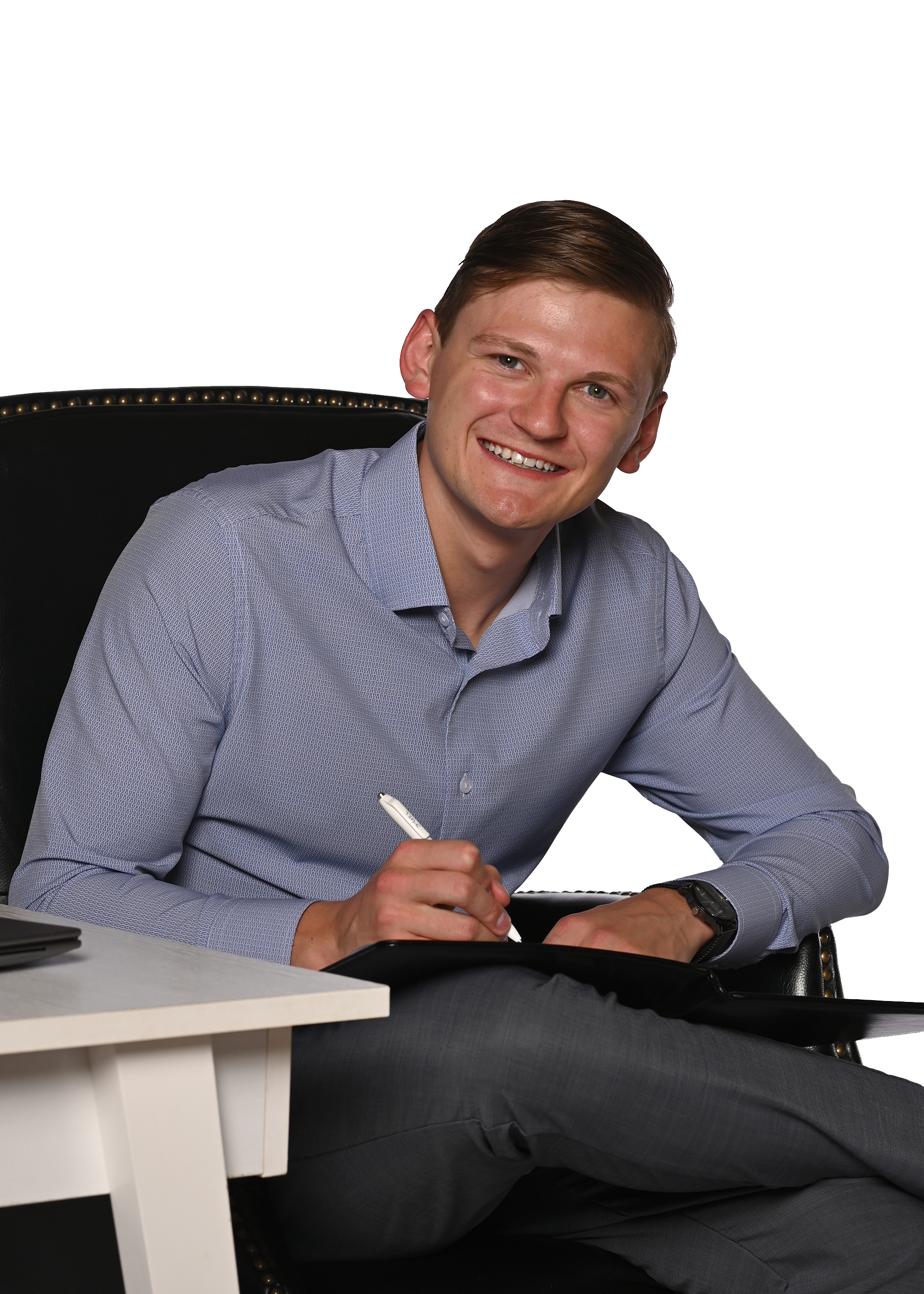 Devin Carver sitting at a desk in a modern office, smiling, wearing a light blue dress shirt, holding a pen, with a black watch on his left wrist.