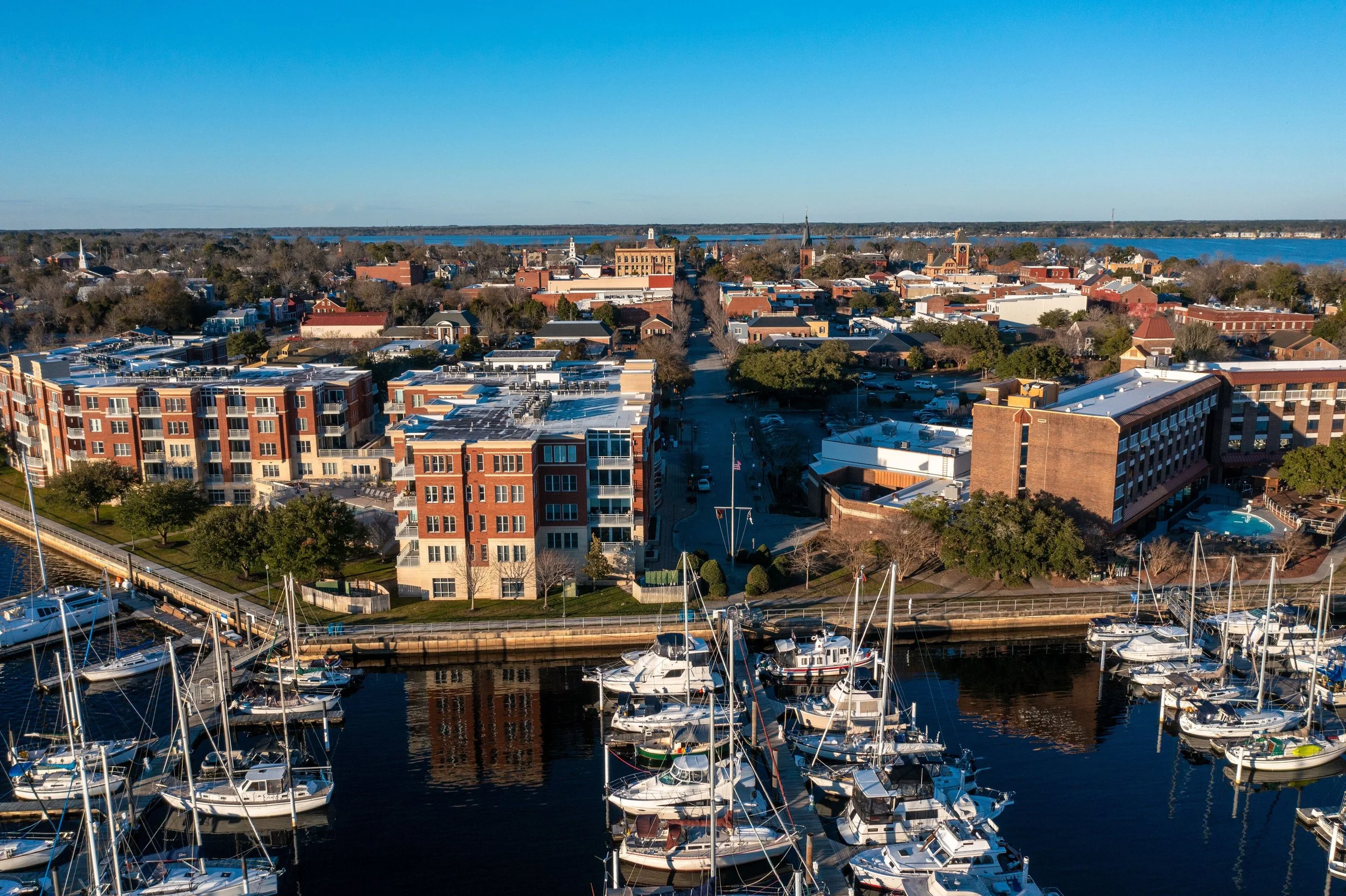 Aerial view of a harbor with sailboats docked, residential and commercial buildings, and trees, with a large river in the background on a clear day.